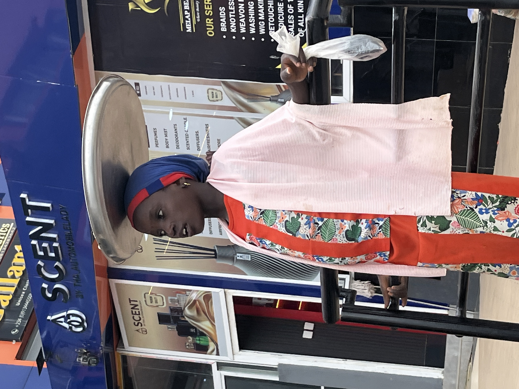 In Galadimawa Village, Nigeria, a young girl is pictured outdoors, balancing a large, round metal tray on her head. She is wearing a red dress with a floral pattern and a light pink shawl draped over her shoulders. A blue and red headscarf is also visible. In her right hand, she holds a white plastic bag. The background reveals the storefront of a business named "SCENT," with advertisements for perfumes, body mist, deodorants, and scented candles. To the right, another advertisement displays "MELAP BEAUTY" and lists services such as braids, knotless extensions, weaving, washing, wig making, retouching, manicures, pedicures, and sales of all kinds. The scene is set in an urban environment with a black metal railing in the foreground.