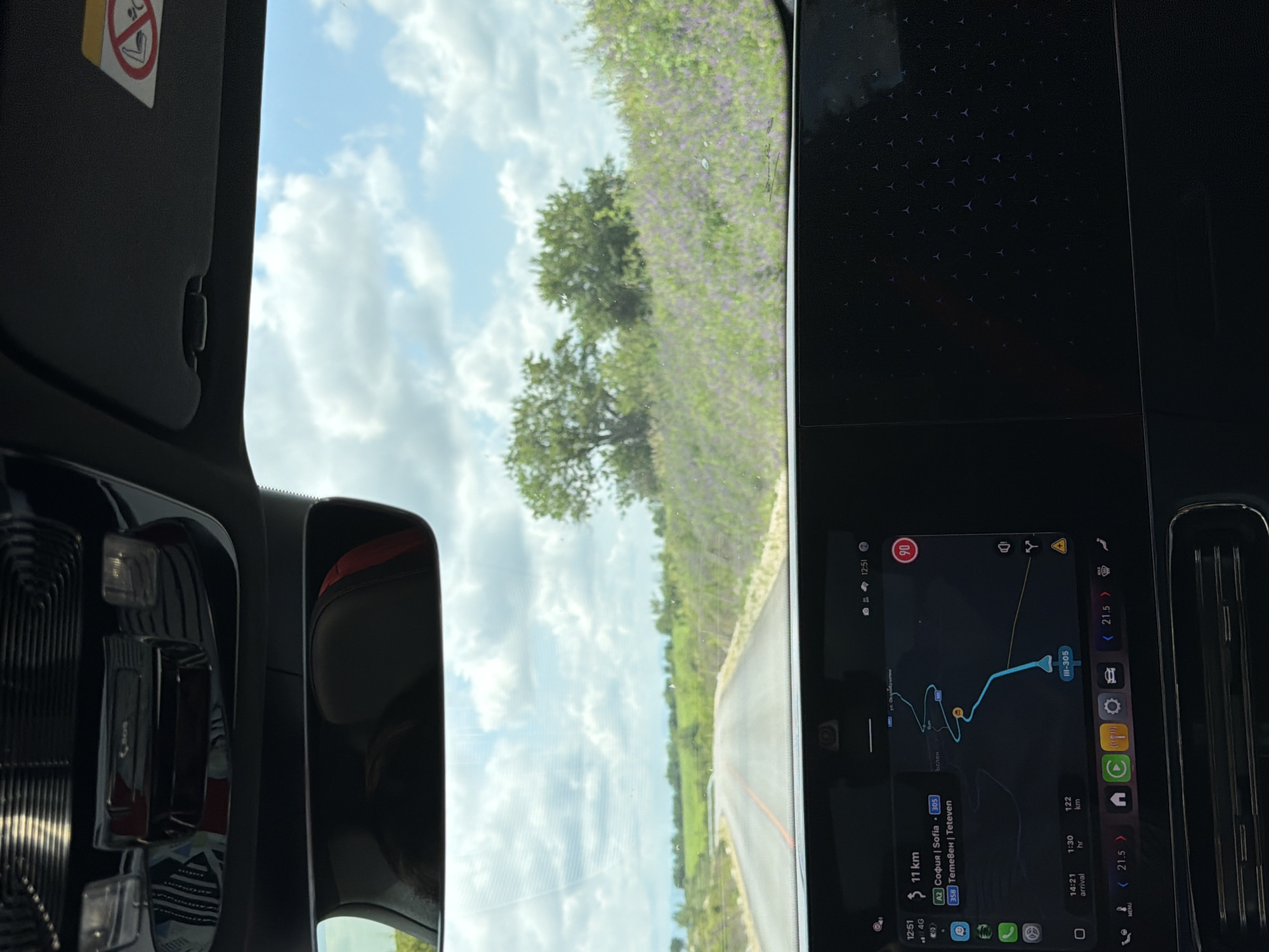 The image captures a view from the interior of a car, looking out through the windshield at a road and rural landscape, while also showcasing the car's active navigation system.
Inside the vehicle, a black sun visor with a red and white airbag warning sticker is visible on the left side of the frame. Part of the car's rearview mirror is also seen. The right side of the image is dominated by a large, dark infotainment screen displaying a navigation map. Adjacent to the screen is a dark panel with a subtle, repeating geometric pattern, possibly part of the dashboard or interior trim, and a car air vent.
Outside the car, a clear blue sky with scattered white clouds is visible. The car appears to be driving on a paved road, which curves slightly to the left. Alongside the road, there are fields with green vegetation and patches of purple flowers, possibly lavender, and a few scattered trees.
The navigation screen shows a route from Sofia to Teteven in Bulgaria, confirming the location hint of Sadovets, Bulgaria, as these are Bulgarian cities and the A2 is a major Bulgarian highway. The current time is displayed as 12:51, with an estimated arrival time of 14:21, indicating a remaining journey time of 1 hour and 30 minutes, covering a total of 122 km. The next segment of the journey is 11 km long. Road numbers like "A2," "358," and "III-305" are visible on the map, along with the name "Ъглен," likely a village or junction. A speed limit of "90" km/h is indicated in a red circle. Other icons suggest mobile signal (4G), Wi-Fi, battery status, and various vehicle settings. The overall impression is of a midday journey through a scenic, possibly agricultural, area.