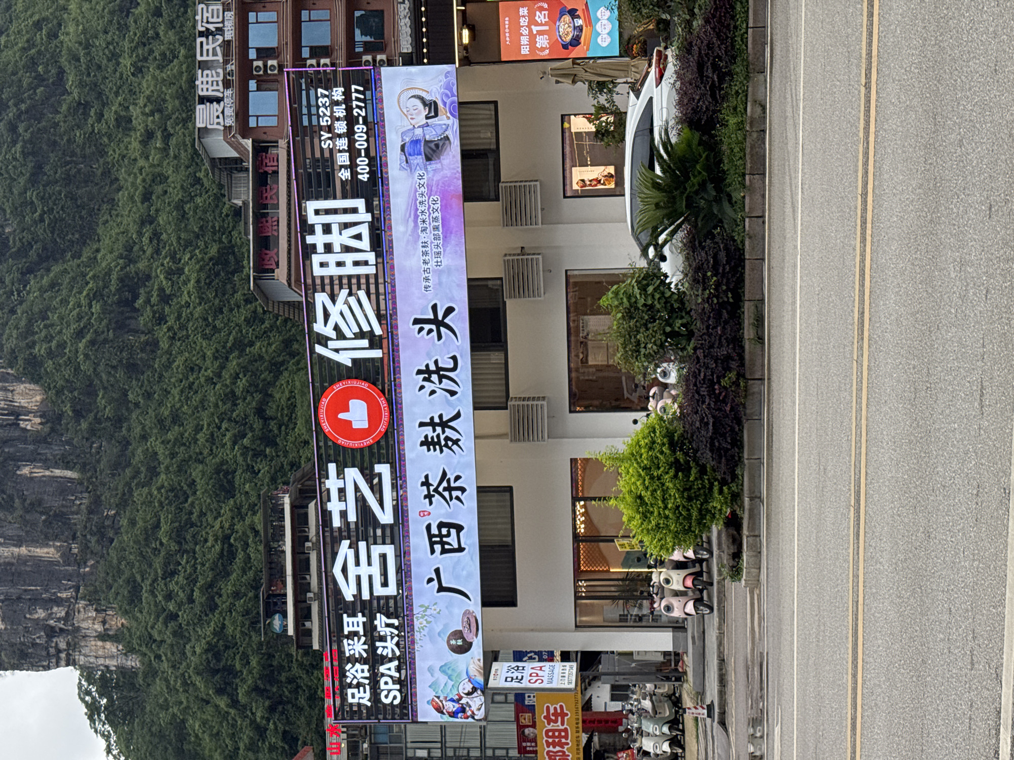 The image shows a commercial street in Guilin, China, likely near a scenic area, given the mountain range visible to the left. A prominent sign advertises a foot massage and SPA business, written in Chinese with a red logo. Adjacent to the sign is another sign advertising "Guangxi Tea Bran Hair Wash," highlighting traditional hair care practices. A three-story building behind the signs appears to be a commercial or residential structure. Numerous parked electric scooters line the sidewalk. A street with double yellow lines separates the sidewalk from the background. The overall impression is of a street scene in a Chinese tourist town.