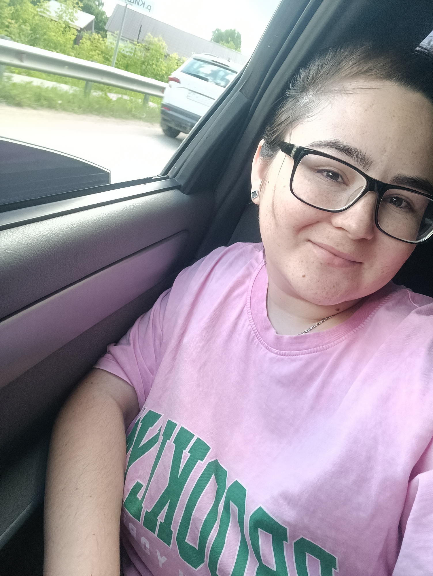 A young Caucasian woman with dark hair, fair skin, and freckles is taking a selfie in a car. She is wearing black-rimmed glasses and a pink t-shirt with green lettering that reads "BROOKLYN." She has a silver chain necklace on. She is smiling and looking at the camera. The background shows the interior of the car, the window, and a blurry view of the outside with a road, a guardrail, trees, and a white car. The image appears to have been taken on a cloudy day.