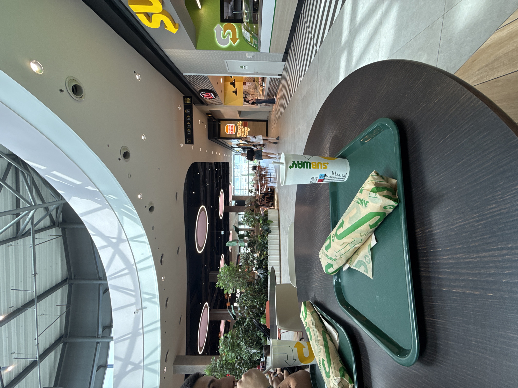 The image depicts a close-up view of a table in a food court, likely in the city of Burgas, Bulgaria, given its association with the "Burgas Plaza" mall. In the foreground, two Subway sandwiches wrapped in branded paper sit on green trays. A Subway branded cup with a straw is also present on one of the trays. The table is dark wood with a visible grain.
In the mid-ground and background, the food court environment is visible. To the right, a Subway restaurant storefront with its signature green and yellow branding is prominent. Adjacent to it is another food establishment, "Burger King," also with its recognizable signage. People are seen walking through the food court and seated at tables in the distance. The interior features a modern architectural design with a large, curved ceiling, artificial lighting, and abundant greenery, suggesting an indoor mall setting. The overall atmosphere appears to be that of a casual dining area within a shopping complex.