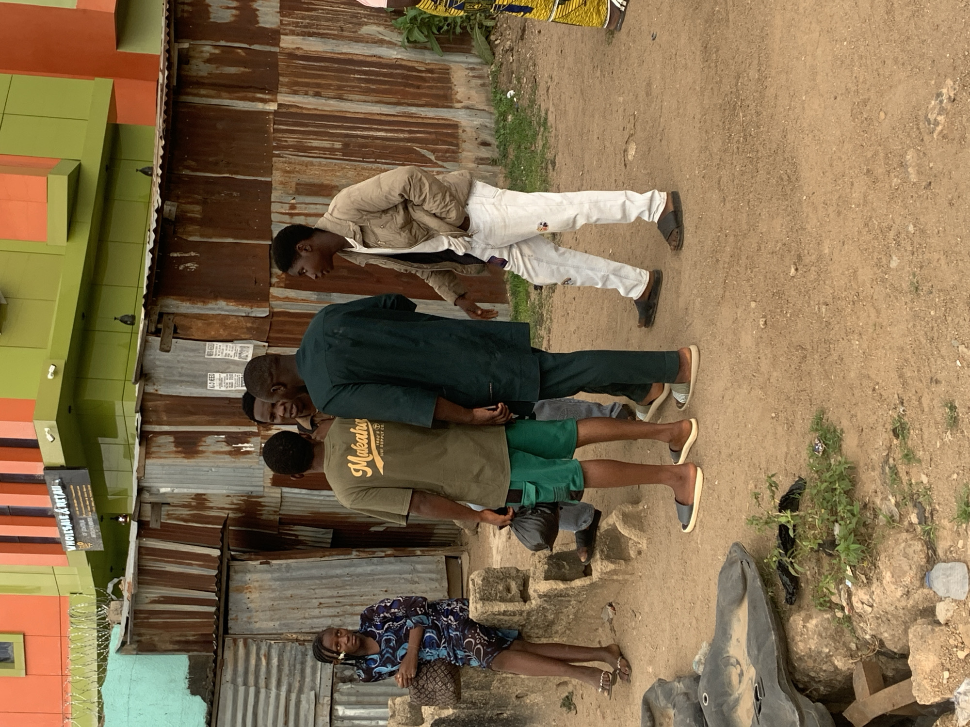 The image shows a group of people standing near a building in an urban area.
**People:**
* Four men are standing together. One is wearing a khaki jacket and white pants. One is wearing a green suit. One is wearing a light green t-shirt with "Makeahu" written on it and green shorts.
* A woman is lying down near a stone structure. She is wearing a blue and black patterned dress.
**Environment:**
* The building behind the group is made of corrugated metal.
* The ground is dirt.
* There is a brightly colored building in the background, painted in green and orange.
**Objects:**
* A black plastic bag.
* A large rock sculpture is visible in the foreground.
* A sign with "WHOLESALE & RETAIL" is seen.
**Location:**
The image appears to be taken in an urban area, given the presence of buildings and people. While the exact location within Jos, Nigeria, cannot be determined from the image alone, the architecture and the general environment suggest a residential or commercial area.