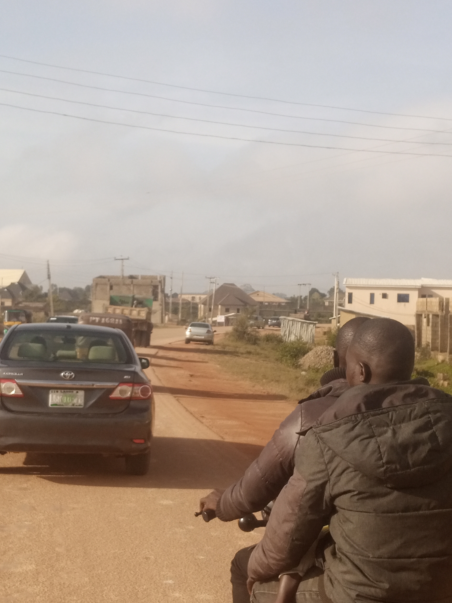 This is a medium shot, eye-level, vertical, outdoor photograph taken in Kwang, Nigeria. The image depicts a dusty, unpaved road with several vehicles and buildings in the background. In the foreground, two individuals are seated on a motorcycle, facing away from the camera. The person in the rear, who is the primary subject, is wearing a dark, padded jacket. The person in front, who is the driver, is partially obscured. A dark-colored sedan is stopped on the left side of the road, with its license plate visible. Further down the road, a large, older truck is parked. The background shows a mix of residential and commercial buildings, some appearing partially constructed. Utility poles and power lines are visible against a hazy sky. The overall impression is that of a developing urban or semi-urban area.