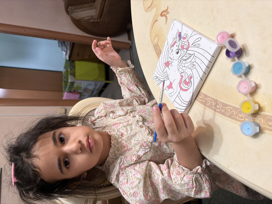 A young female, approximately 5-8 years of age, is depicted seated at a light-colored table in an indoor residential setting in Таскино, Russia. She has dark hair, styled with a pink clip on the top of her head, and dark eyes, looking directly towards the camera. She is wearing a long-sleeved, high-necked blouse with a ruffled collar and cuffs, featuring a light floral pattern of pink and green on an off-white background.
Her right hand holds a blue-handled paintbrush with a red tip, poised over a small white canvas. The canvas displays a "paint by number" outline of a unicorn or mythical creature, with portions of its mane and a small body section already colored pink and purple. To her right, a connected strip of seven small plastic paint pots is arranged on the table, containing various pastel colors including light blue, yellow, light pink, dark blue, purple, and light purple, each marked with a number.
The background reveals a domestic environment. Behind the subject, a brown door frame leads into another room where a light green playpen or crib is visible. To its right, a brown chest of drawers with items on top, and a brown upholstered armchair or sofa, are also present. The lighting is consistent with indoor illumination.