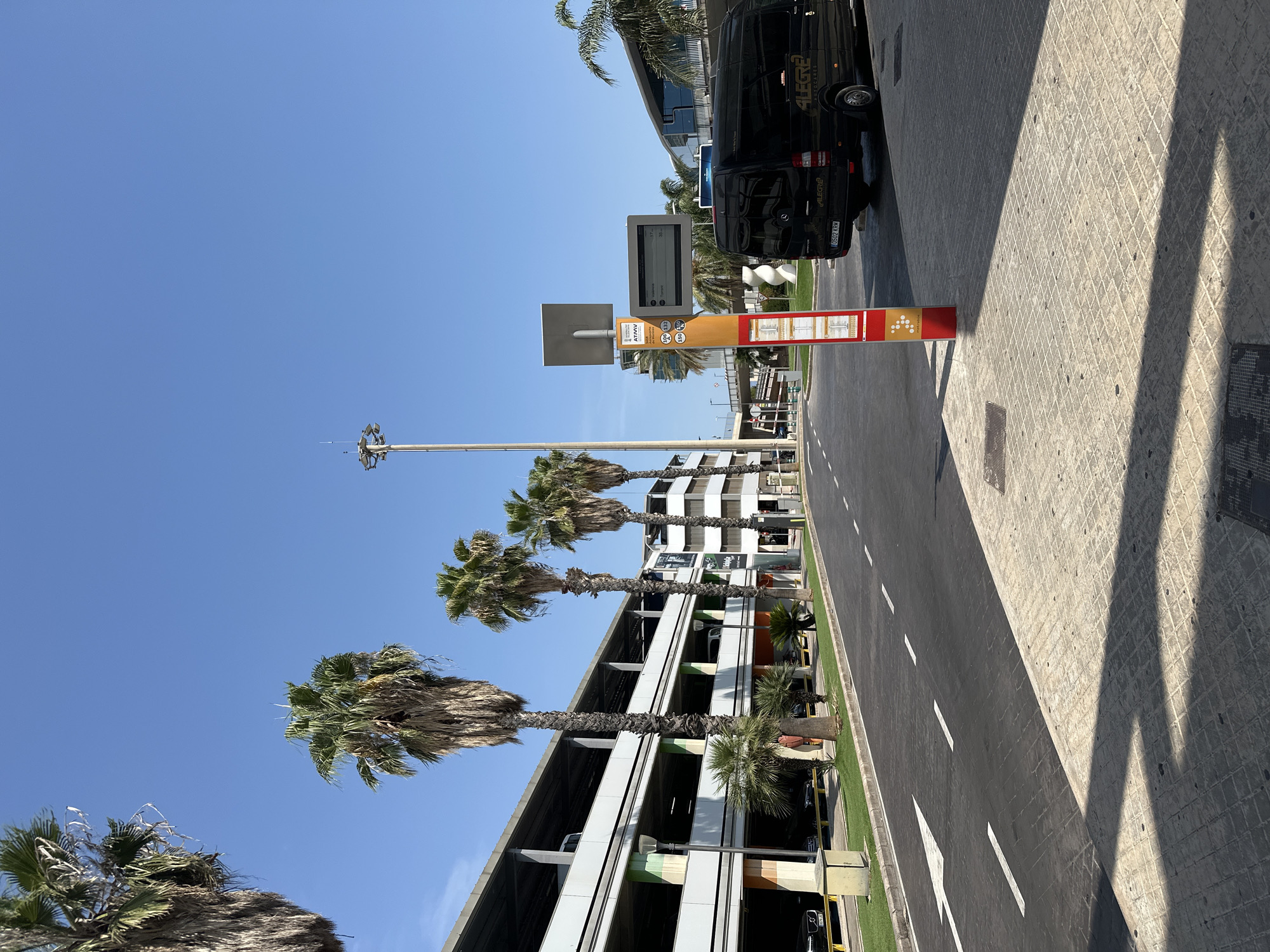 This image depicts an outdoor scene in Manises, Spain, likely near an airport or transportation hub, given the presence of a multi-story parking structure, a bus stop sign with a digital display, and a passenger van. A row of tall palm trees lines a paved road with white lane markings. The parking garage, constructed with concrete levels and supporting columns, is visible on the left, with cars parked on its lower levels.
In the foreground, a red and yellow bus stop pole stands prominently. It features a digital screen displaying arrival times for "Valencia" (17 minutes) and "Torrent" (36 minutes), along with route numbers 150 and 20. Below the digital display are smaller informational panels.
A black passenger van, branded with "ALEGRE AUTOCARES," is parked to the right of the bus stop. The van has a license plate that reads "0502 KRM." The road surface appears to be asphalt with a textured, lighter colored paving in the foreground, casting a distinct shadow pattern. In the background, a large, modern building with a curved roof and glass facade is visible, possibly an airport terminal. A tall light tower with multiple lamps and an antenna is also a significant feature against the clear blue sky. The overall environment suggests a sunny day in a well-maintained urban or transportation infrastructure setting.
