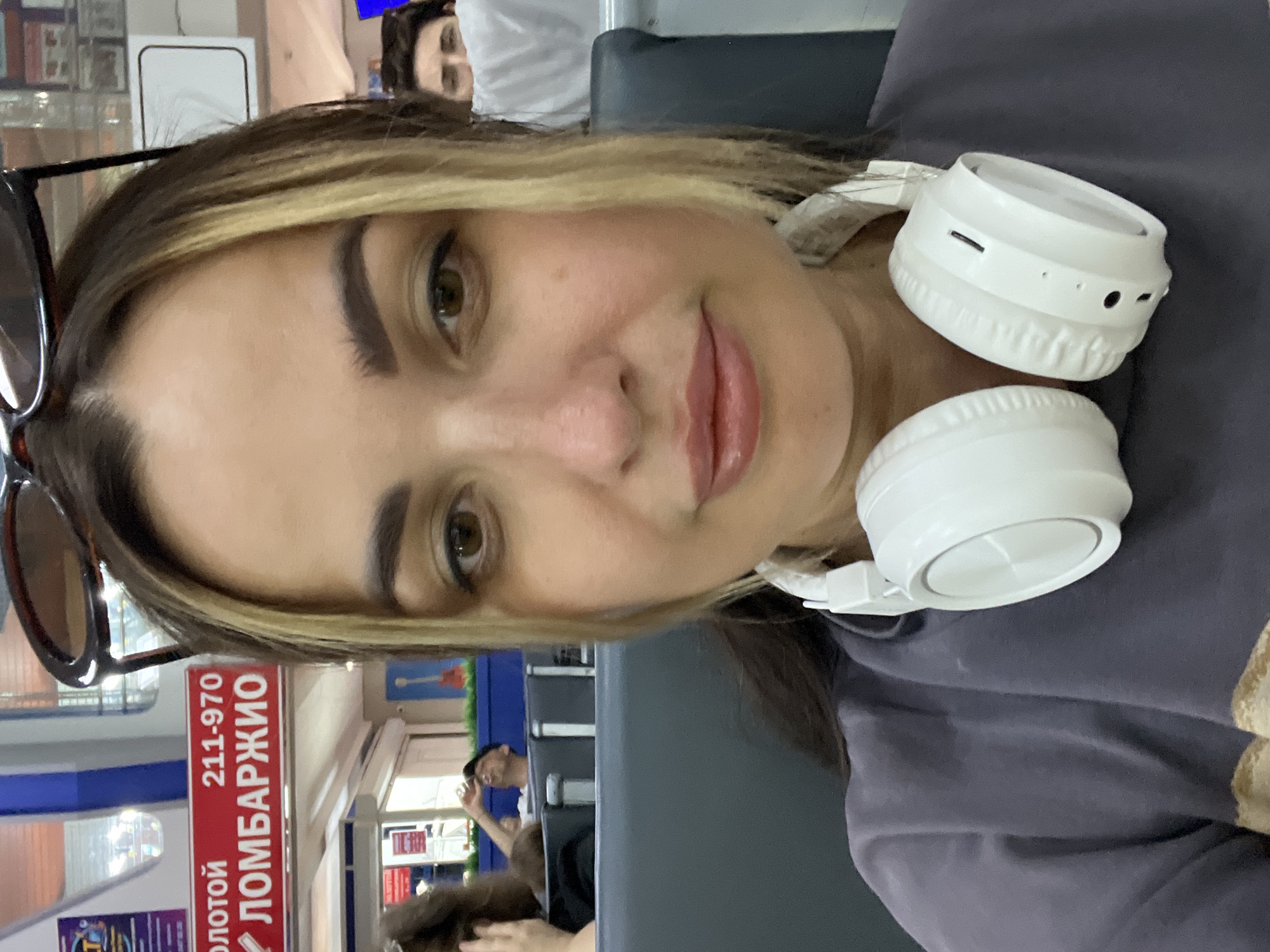 The image shows a close-up, selfie-style shot of a woman with blonde highlights in her dark hair. She is wearing white headphones around her neck and a dark purple t-shirt. Her face is the primary focus, with her eyes looking forward and her lips slightly parted. In the background, out of focus, are other people sitting in what appears to be an airport or waiting area. A red sign with white text is visible in the upper left portion of the image, reading "ЗОЛОТОЙ 211-970 ЛОМБАРДЖИО". The overall lighting suggests an indoor environment, possibly during the day.