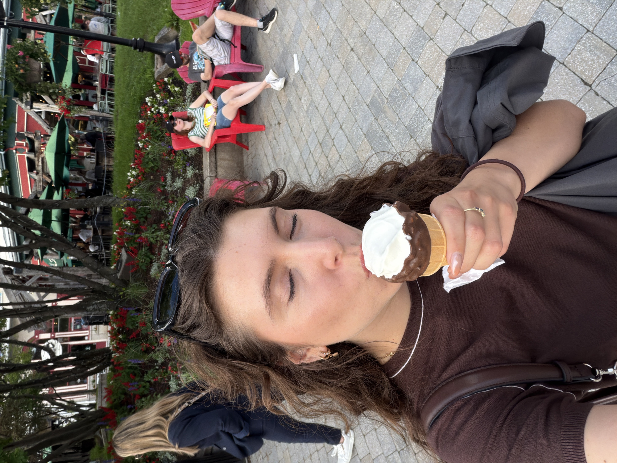A young woman is captured in a close-up selfie, enjoying an ice cream cone outdoors. Her eyes are closed as she takes a bite of the white ice cream with a chocolate-dipped cone, holding it with her right hand. She wears a dark brown short-sleeved shirt with a white trim around the neckline and a dark grey jacket draped over her left arm. A dark purse strap is visible on her left shoulder. A thin gold necklace and a ring with a small dark stone adorn her hand. In the background, the paved ground is visible, leading to a park-like setting with trees, colorful flower beds, and people seated on red chairs under green umbrellas. The scene appears to be in the pedestrian village of Mont-Tremblant, Quebec, Canada, known for its vibrant atmosphere and outdoor seating areas.
