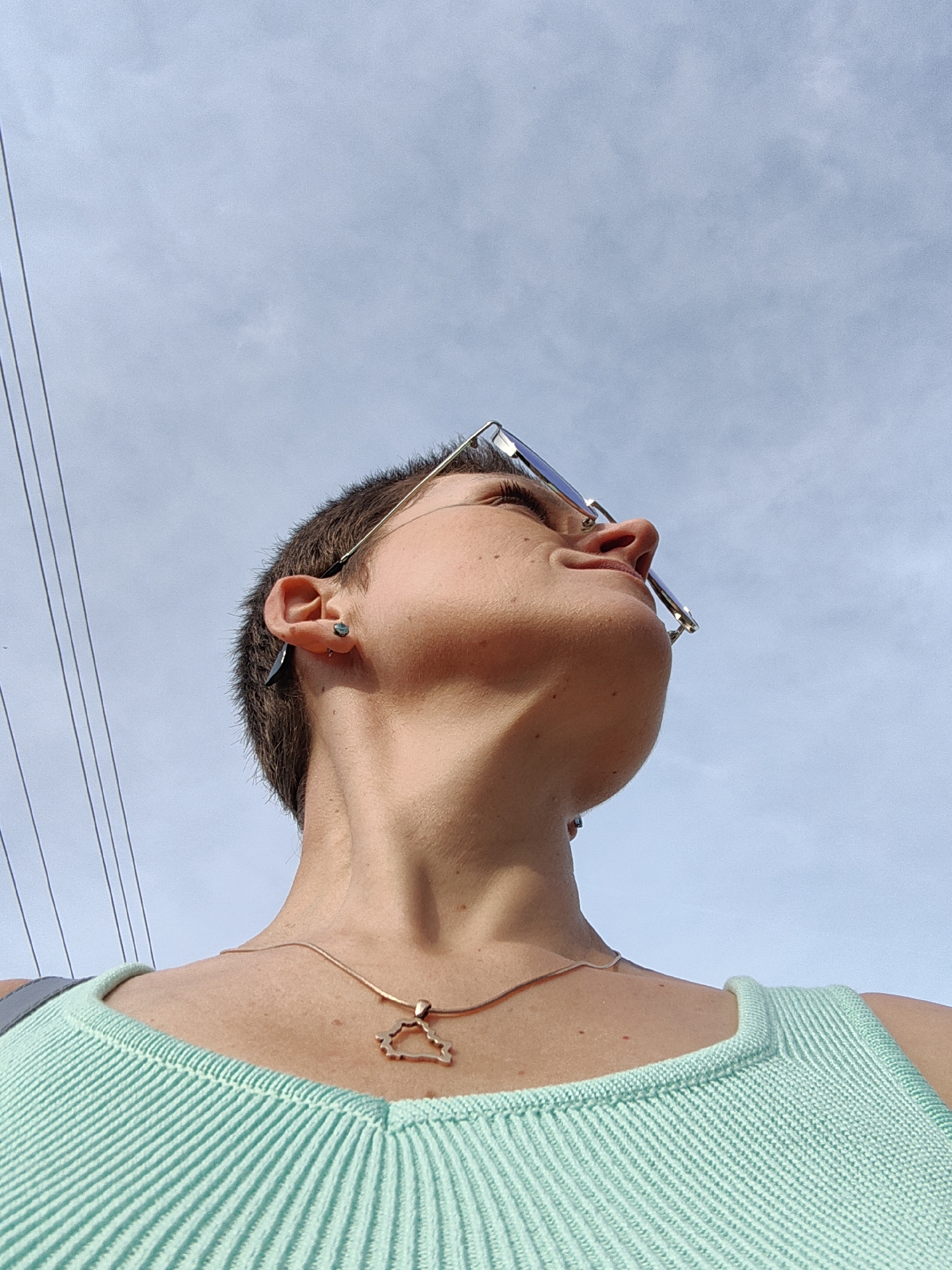 A low-angle, outdoor, waist-up shot shows a person looking upwards towards a cloudy sky. The person has short, dark hair and is wearing a mint green ribbed tank top and a delicate gold necklace with a cloud-shaped pendant. They are also wearing sunglasses with thin, silver frames and have earrings in their left ear. The background is a pale blue sky with scattered clouds and thin, dark wires crisscrossing the upper left portion of the frame. The lighting suggests it is daytime. The image was taken in Fanipal, Belarus.