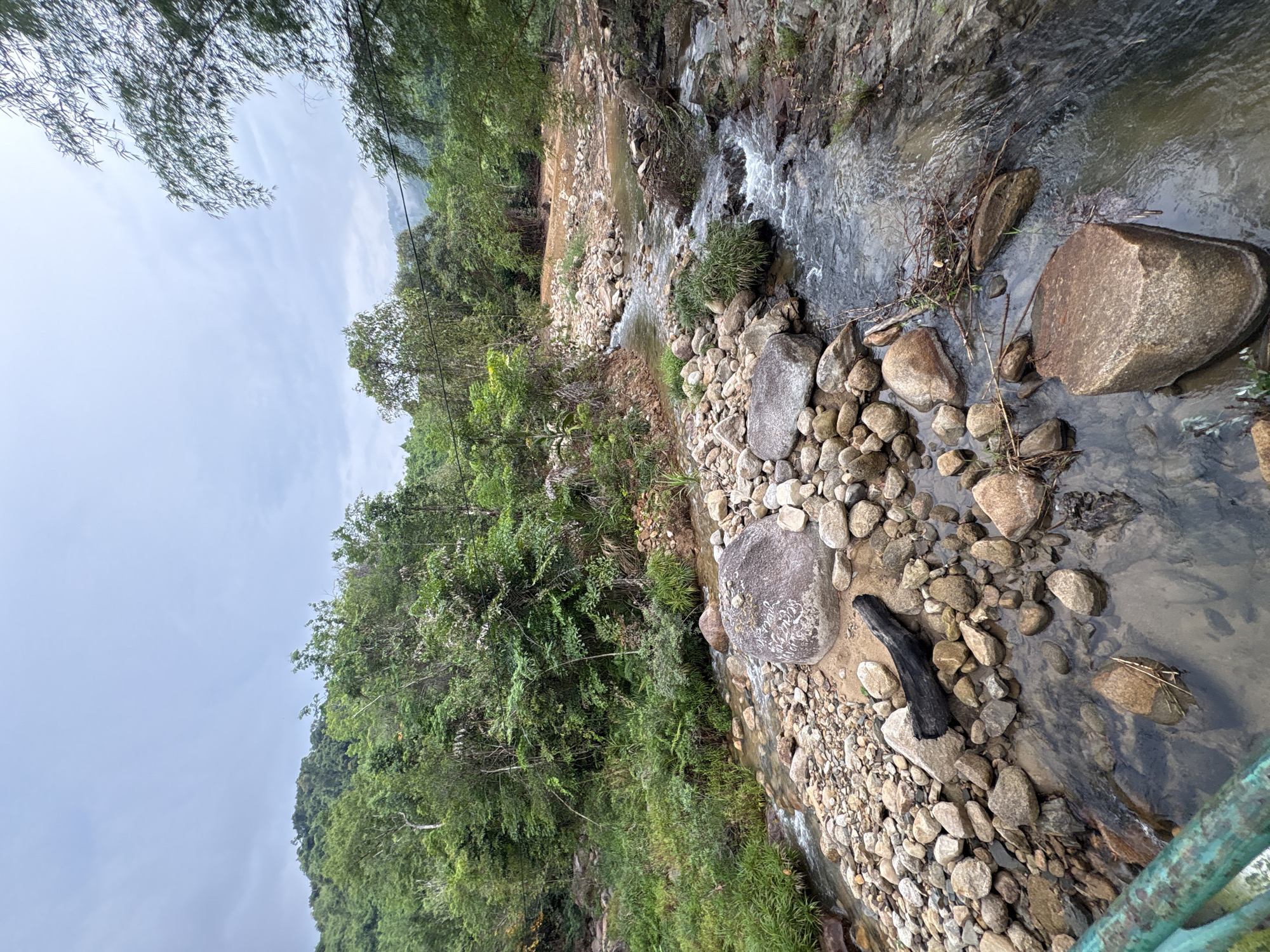 The image depicts a shallow, rocky stream flowing through a terrain in Khanh Vinh, Vietnam. The water flows around and over various sized rocks and pebbles that line the streambed. A piece of dark, possibly charred wood rests among the rocks near the foreground. The stream banks are vegetated with green foliage, which extends up the surrounding hillsides. The sky is overcast, casting a diffused light over the scene. The perspective suggests the view is captured from an elevated position, potentially from a bridge.