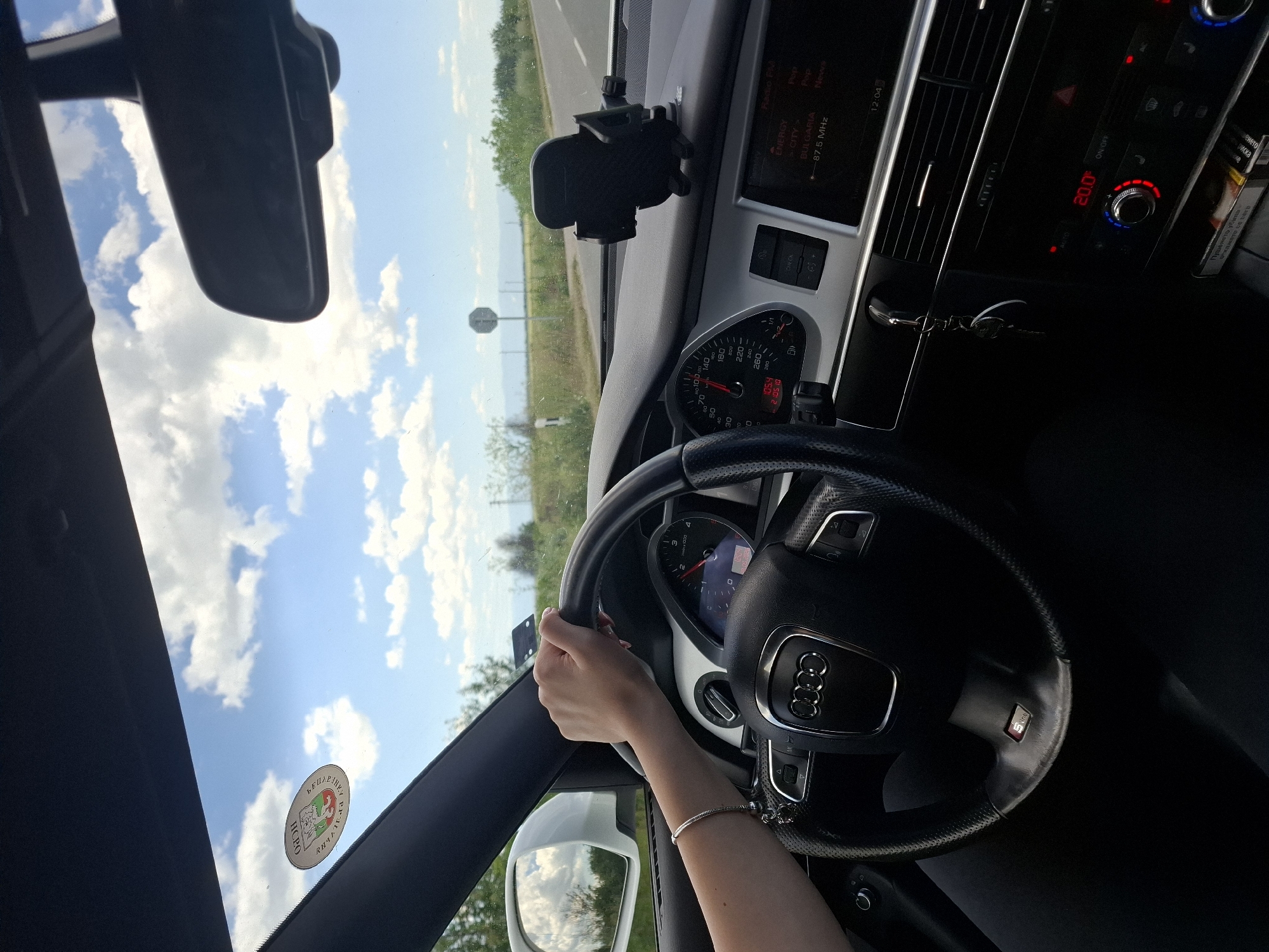 A person is driving an Audi, likely an S-Line model given the steering wheel badge, on a road near Brezovo, Bulgaria. The environment is sunny with a blue sky and scattered clouds. The car's interior features include a dashboard displaying a temperature of 20.0 degrees Celsius, radio tuning to 87.5 MHz (Bulgaria), a navigation system (or infotainment screen), and a cell phone holder mounted on the dashboard. A sticker, likely related to a local municipality, is visible on the windshield near the rearview mirror. The driver's hand is on the steering wheel, adorned with a bracelet. The speedometer indicates a speed that is hard to distinguish, and the tachometer shows low RPM.