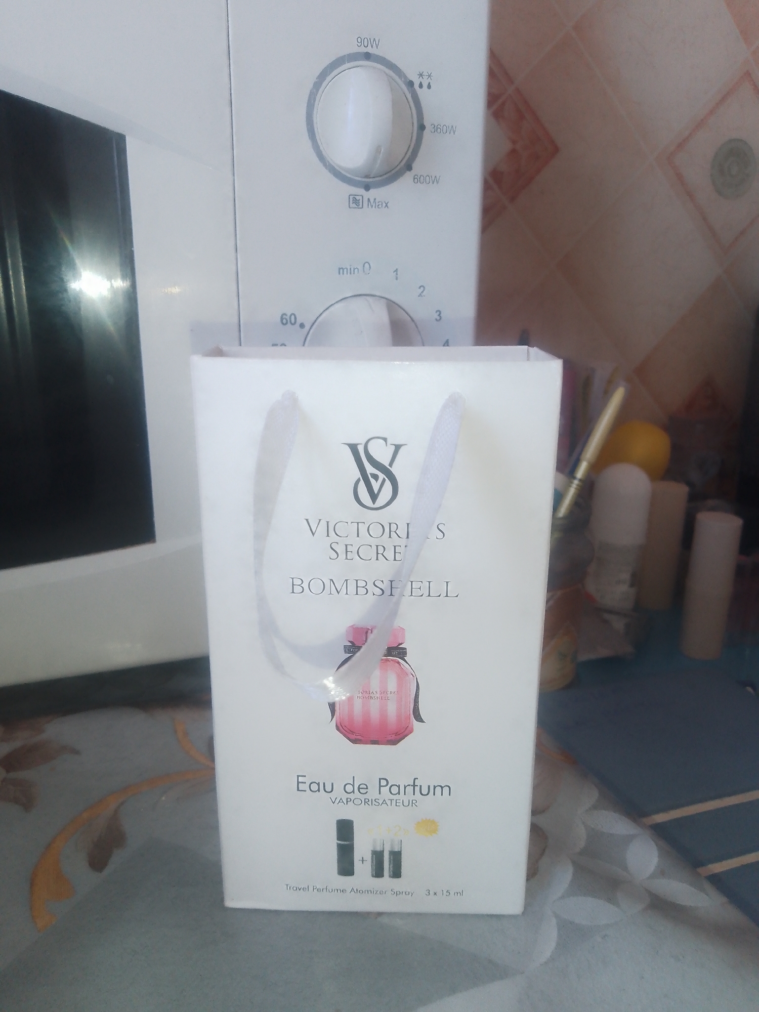 The image shows a white paper gift bag with the Victoria's Secret logo and the text "Bombshell Eau de Parfum Vaporisateur Travel Perfume Atomizer Spray 3 x 15 ml". The bag is placed on a surface in front of a white microwave oven. The background shows a tiled wall and a collection of items on a shelf, including a yellow object, a roll-on deodorant, and two cylindrical objects that appear to be makeup removers or applicators. The scene is indoors, likely in a domestic setting. The image does not provide any specific location information that would place it in Chernivtsi, Ukraine.