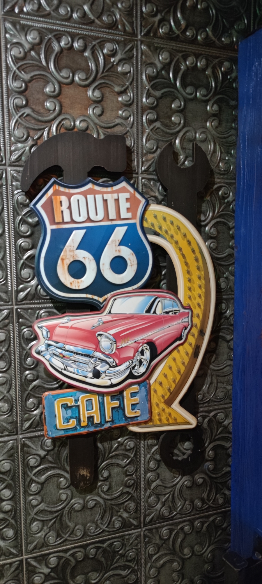 This is a close-up, vertical shot of a decorative metal sign, likely a wall decoration, placed against a dark, textured wall with a repeating embossed pattern of swirling filigree. The sign itself is shaped and decorated to evoke a retro, automotive theme. Dominating the upper portion of the sign is a shield-shaped emblem with a brown and white border, featuring the words "ROUTE 66" in prominent white lettering against a blue background. Below this is a stylized depiction of a classic red car, possibly a 1950s model, with chrome accents and whitewall tires. Underneath the car is a rectangular sign with the word "CAFE" in bold, yellow letters against a blue and orange background. The entire sign assembly appears to be made of metal and is layered, with some elements appearing to be raised or embossed. To the left of the Route 66 emblem, the silhouette of a hammer is visible, and to the right, the silhouette of a wrench is present, further reinforcing the automotive and workshop theme. The overall aesthetic suggests a diner, bar, or garage setting with a nostalgic American flair. There are no people visible in the image. The lighting is moderate, highlighting the textures and colors of the sign. The background wall has a subtle sheen, indicating it might be a metallic panel or a very dark, glossy paint. The right edge of the image shows a sliver of a bright blue wooden surface, likely part of a door or another wall element, providing a contrasting color to the dark tones of the sign and background. There is no specific indication of the location being in Yerevan, Armenia, as the content is universally themed and doesn't contain any identifiable Armenian landmarks or text.