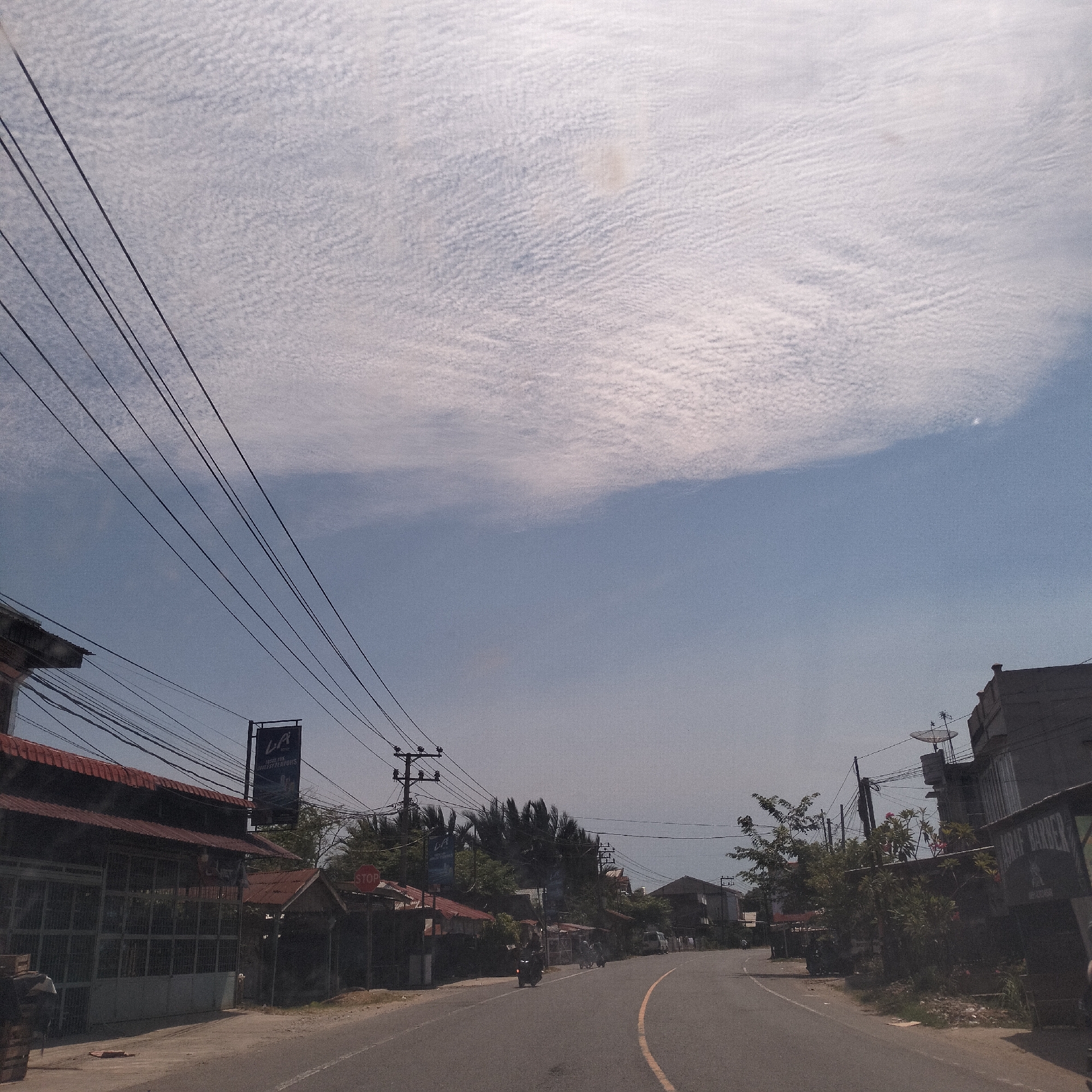 The image depicts a street scene in Cibrektunong, Indonesia, on a day with thin, high clouds filling the sky. A two-lane road with a yellow center line curves slightly to the right. The road is flanked by buildings and vegetation. The buildings appear to be a mix of commercial and residential structures, some with red tile roofs. Power lines crisscross overhead. A sign is visible on the left side of the road, bearing the letters "LA" above smaller text. A stop sign is also visible. A few motorcycles can be seen on the road. The overall impression is of a typical street view in a small Indonesian town on a sunny day.