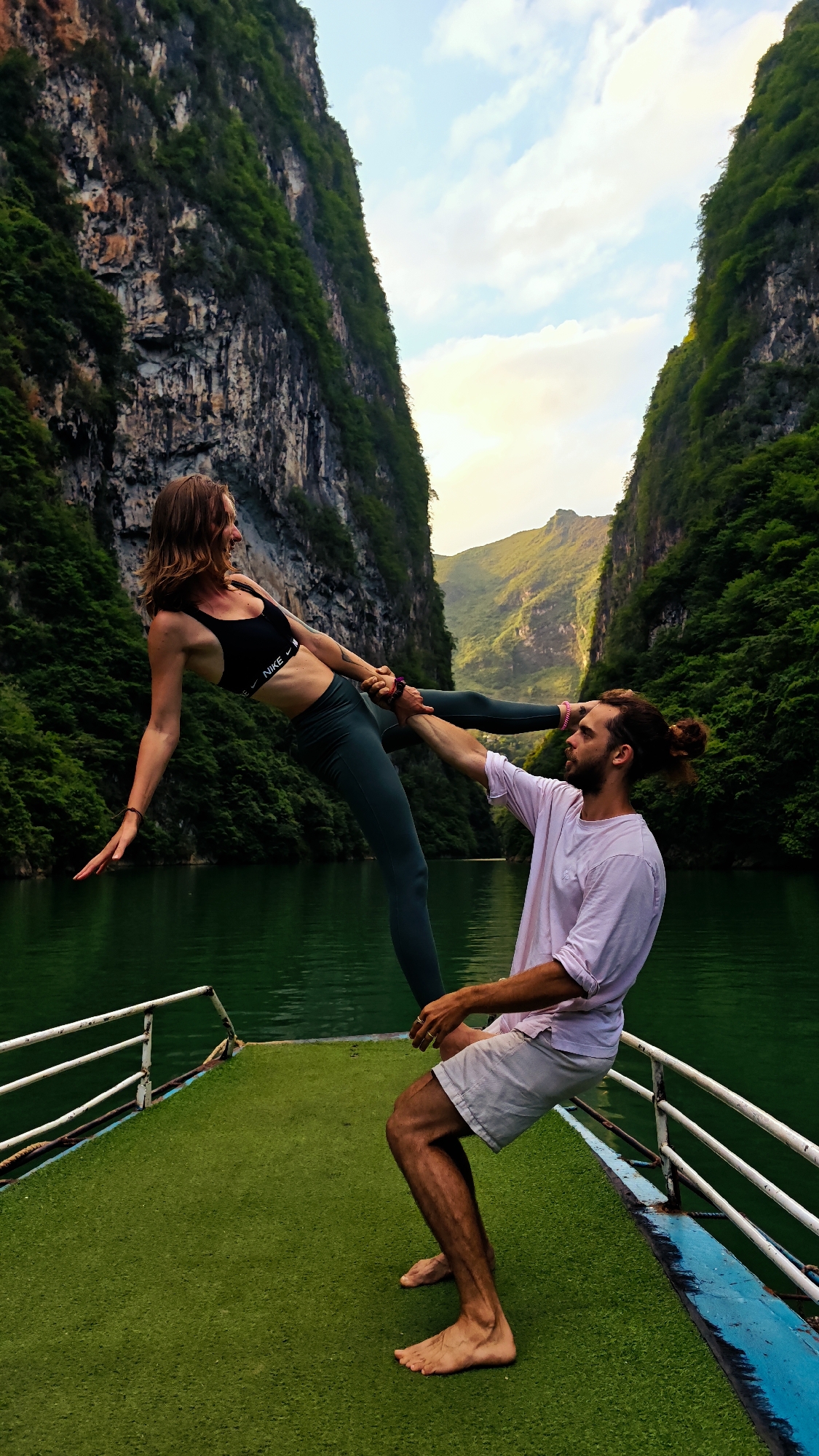 On a boat within the Đồng Văn Commune in Vietnam, a woman is performing a yoga pose supported by a man. The woman, wearing a black sports bra and dark green leggings, is balancing horizontally, holding the man’s hands. The man, wearing a light pink shirt and grey shorts, is bent over supporting her. They are on a boat with a green artificial turf deck and white railings. The river water is a deep green, flanked by steep, rocky cliffs covered in green vegetation. In the background, mountains are visible under a partly cloudy sky.