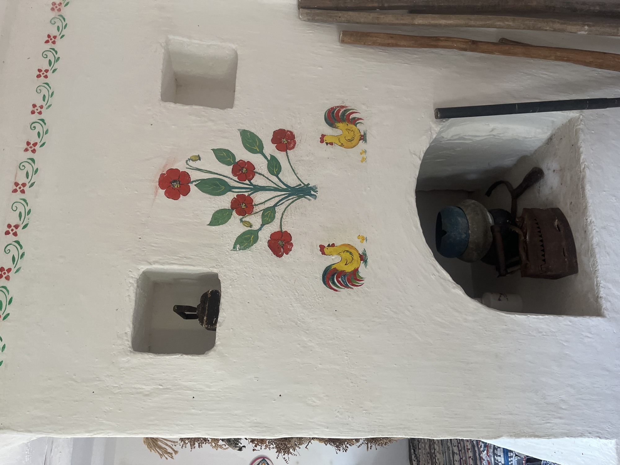 The image shows the interior of a traditional Russian stove, likely located in Taman', Russia. The stove is whitewashed and decorated with painted motifs of red flowers and green leaves along the top border. In the center of the stove, a bouquet of red flowers is painted. To the left and right of the flower bouquet are painted roosters with chicks.
There are two recessed niches in the stove. The upper left niche contains a dark, metallic object with a handle. The upper right niche is empty.
Below the painted decorations, there is a large arched opening that serves as the hearth of the stove. Inside the hearth, on a shelf, are several objects: a blue and black round object with a handle, an old-fashioned iron, and other unidentifiable metallic items. To the right of the hearth, leaning against the stove, are three wooden poles or sticks.
The wall to the far left of the image shows a portion of a rug with a geometric pattern and what appears to be dried plants hanging from the ceiling. The overall impression is one of a rustic, historical setting, possibly a museum exhibit or a preserved traditional home.