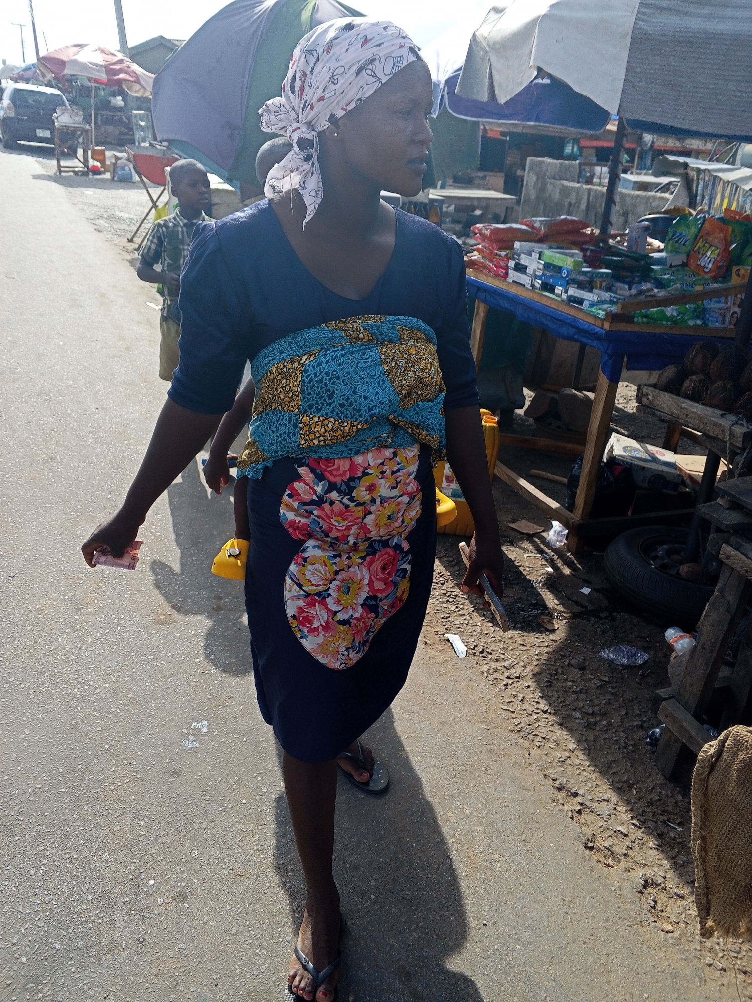 This full-shot image captures a vibrant street scene, likely in a market area in Adankolo, Nigeria, during bright daylight. The primary subject is a woman walking towards the right side of the frame, her gaze directed forward. She has dark skin and is wearing a dark blue dress with a v-neck and short sleeves. A colorful blue, yellow, and black patterned fabric is wrapped around her midsection, with a vibrant floral fabric (red, pink, yellow, blue flowers) visible below it, indicating a child carried on her back. A yellow shoe of the child is faintly visible. The woman's head is covered with a white headscarf adorned with a light pink pattern, tied neatly on the side. She wears simple dark flip-flops. In her left hand, she holds several pink banknotes, and in her right, a silver-colored object, possibly a phone or small tool.
Behind the woman, to her left, a young boy in a plaid shirt and light shorts is visible, also looking towards the right. The background features a bustling roadside market with several makeshift stalls. To the right, a stall with a blue tarp displays various packaged goods like snacks and other small items on a wooden table, with coconuts piled nearby. Further into the background, other stalls are covered by large umbrellas, including one with red and white stripes and others in green and dark blue, providing shade. A dark-colored vehicle is partially seen in the far left background. The woman and boy are walking on a paved road, while the ground alongside the market stalls is uneven, dusty, and strewn with some debris. Strong shadows on the ground indicate bright sunlight. No clearly legible text is visible on the market goods or elsewhere.