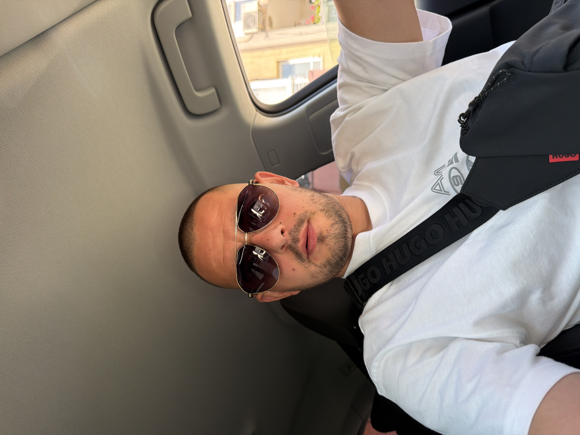 A man with a shaved head and stubble is wearing aviator sunglasses, a white t-shirt, and a black bag with a strap that says "HUGO BOSS". He is sitting in the driver's seat of a car, with a seatbelt visible. The interior of the car is visible, as well as the exterior through the window, showing buildings that appear to be in the city of Burgas, Bulgaria. The lighting suggests it is daytime.