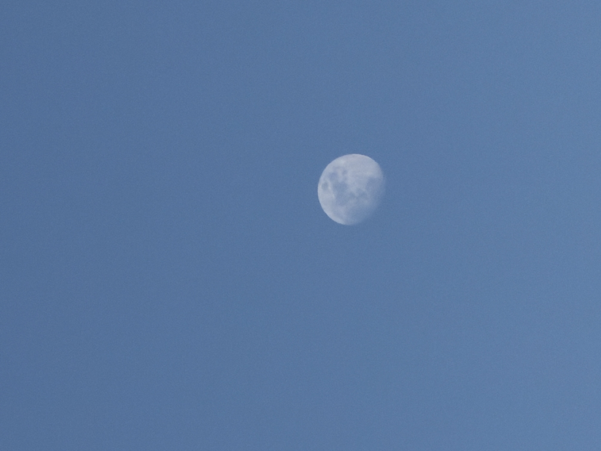 The image features a prominent, roughly spherical moon against a clear, uniform medium-blue sky. The moon, appearing in a gibbous phase, is positioned slightly right of the center of the frame. Its surface exhibits visible greyish and white textures, suggesting craters and lunar maria, with subtle shadow details indicating its three-dimensional form.
The scene depicts the moon visible during the daytime, commonly referred to as a "day moon," as evidenced by the bright, cloudless blue sky. There are no clouds, sun, stars, or other celestial bodies visible, creating a minimalist composition focused solely on the moon and the vast expanse of the sky. No people, man-made objects, or vegetation are present in the frame. The weather appears clear and sunny. No specific activity or event is taking place, only the passive observation of the moon. There is no visible text. The location is stated to be Sofia, Bulgaria.