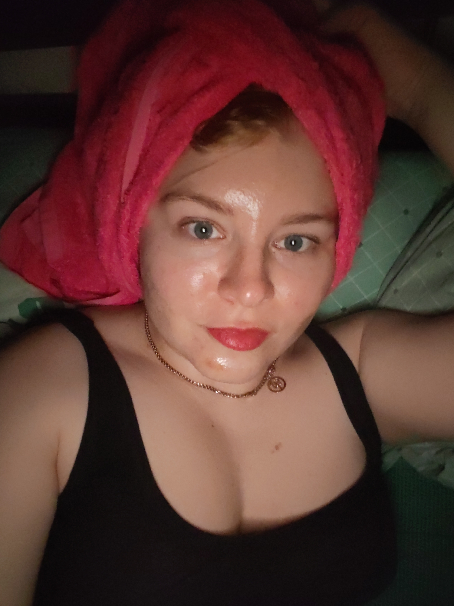 A fair-skinned woman with blue eyes is lying down. She is wearing a black tank top and a gold necklace with a circular pendant. Her hair is wrapped in a pink towel. The background appears to be a green and white patterned pillow or blanket. There is no information available to determine if this photo was taken in Izhevsk, Russia or any other location.