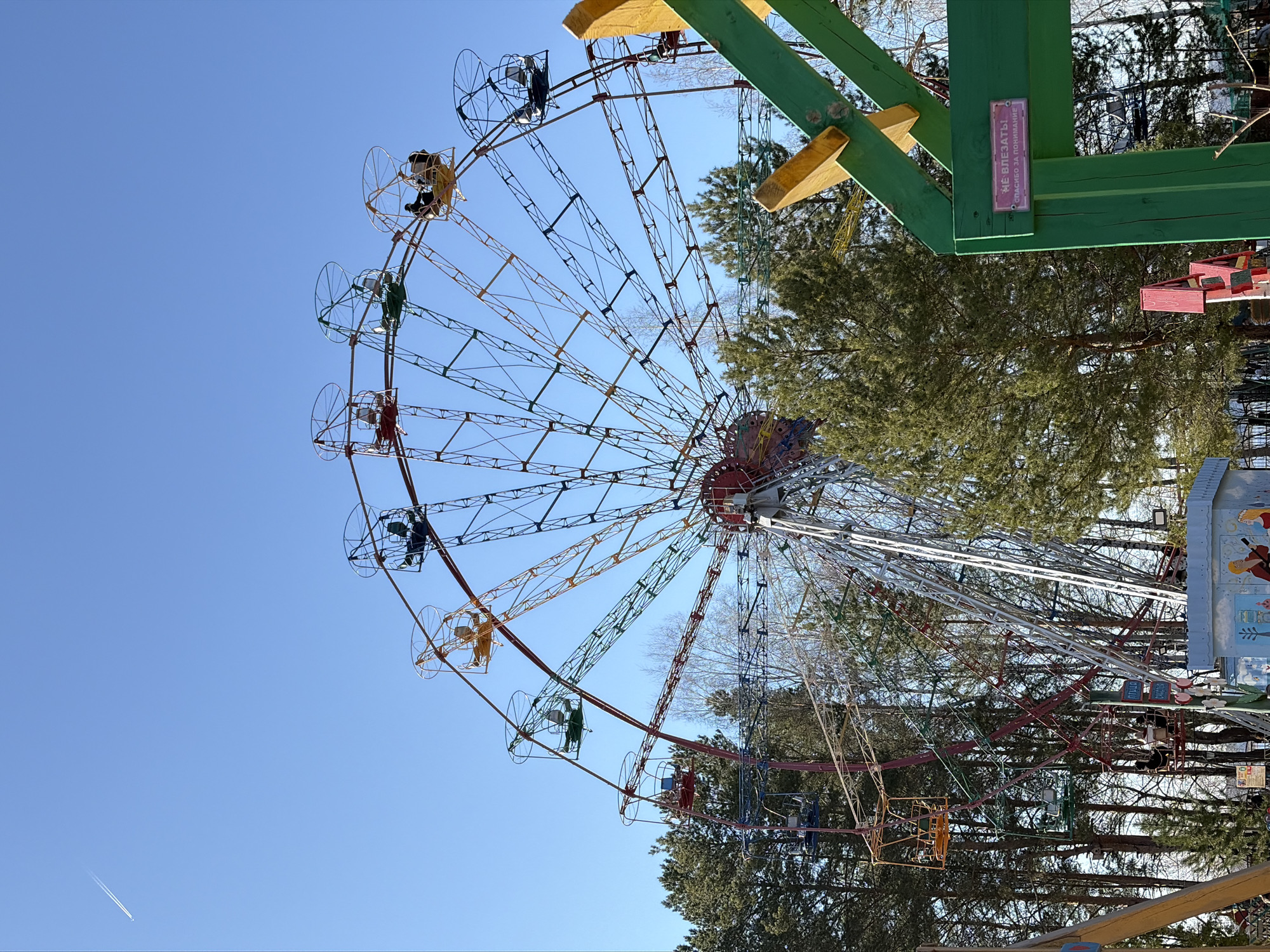 A Ferris wheel, located in the city of Chaikovsky, Russia, dominates the scene. The Ferris wheel is characterized by multi-colored carriages suspended from a network of steel beams. Several occupants are visible in the carriages. The structure is surrounded by trees and a clear blue sky. A sign in Russian is attached to a wooden support structure.