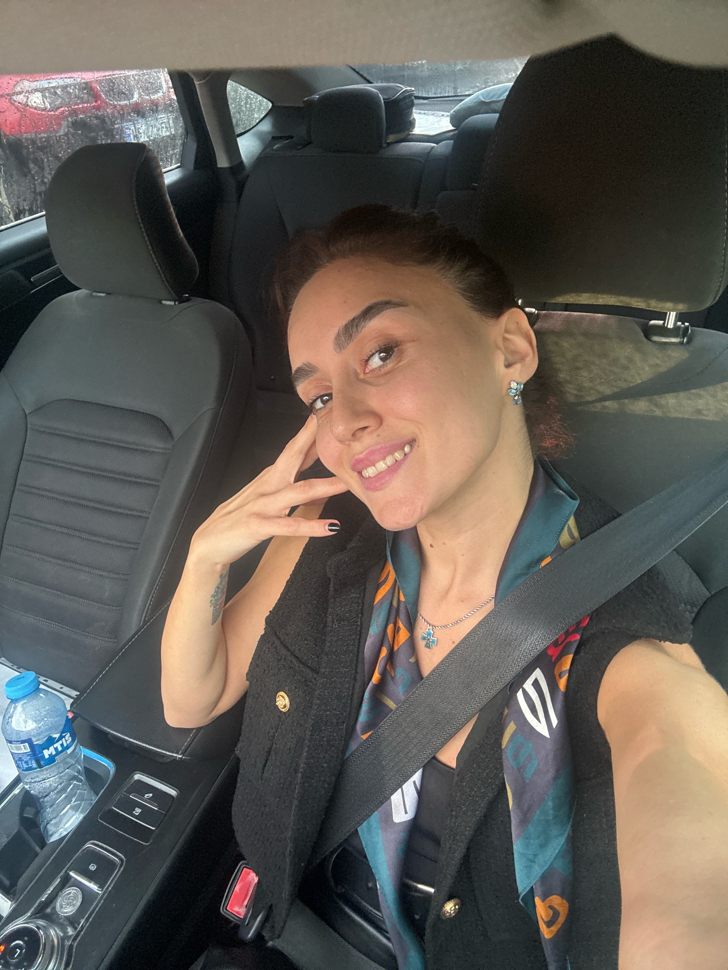 A woman with dark hair and brown eyes is taking a selfie in the driver's seat of a car. She is wearing a black textured jacket over a black top and a colorful patterned scarf. She has on a silver necklace with a blue cross pendant and blue floral earrings. Her right hand is touching her face, and she is smiling at the camera. Outside the car window, it appears to be raining, with other vehicles visible in the background. On the center console, there is a bottle of water and car controls. The interior of the car has black leather seats. The scene is set in the city of Tbilisi, Georgia, indicated by the presence of the water bottle brand "MTIS," a common brand in Georgia.