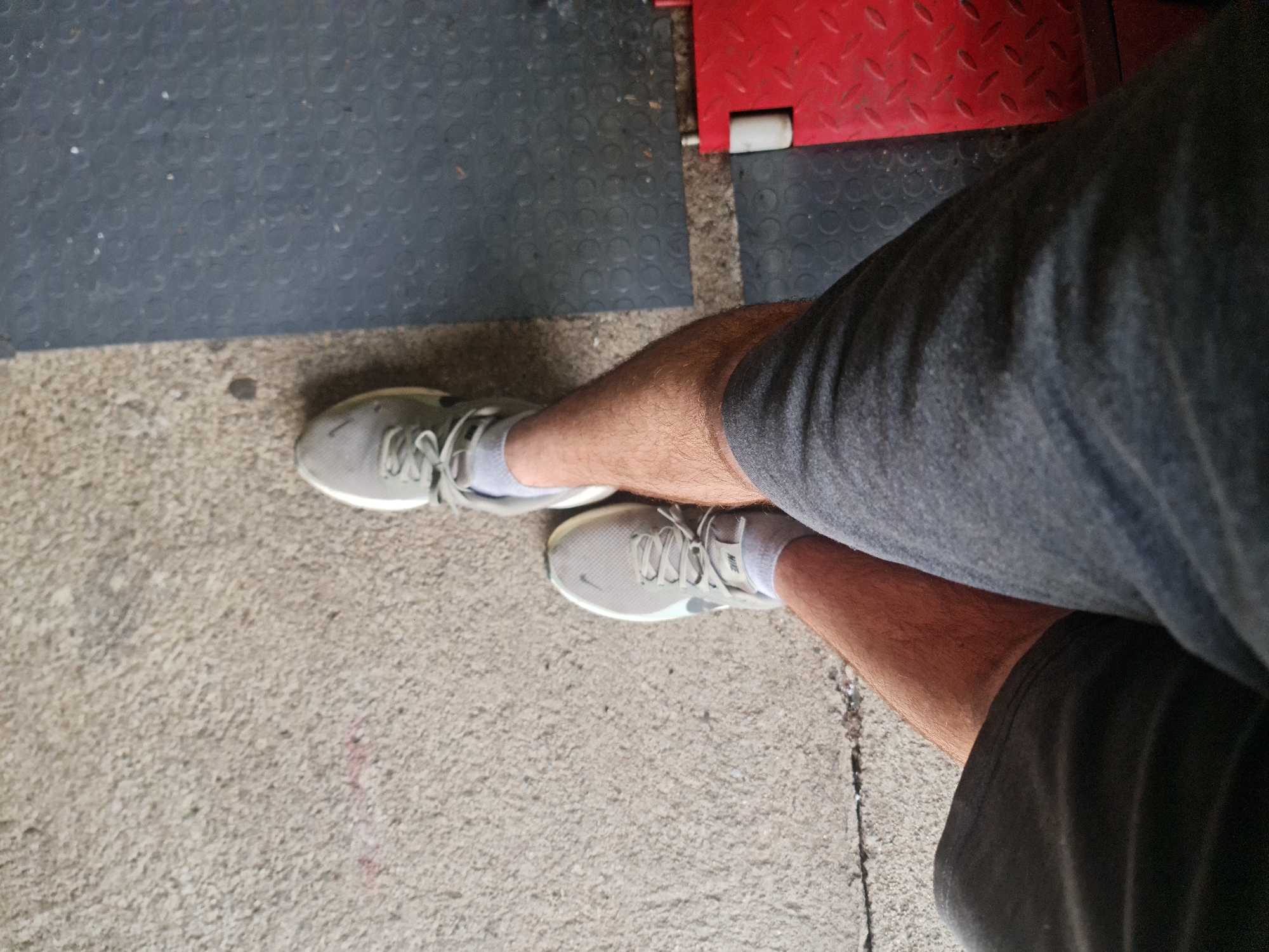 The image, taken from an overhead perspective, captures the lower body of an individual, likely standing or seated. The person wears grey athletic shorts and white, low-cut socks. Their legs are visible, covered in light brown hair. On their feet are a pair of light grey athletic shoes, which appear to be Nike brand, with a faint "NIKE" text visible on the sock above the tongue of the right shoe and a partial swoosh logo on the side of the left shoe.
The setting appears to be an indoor or semi-outdoor functional space. The ground is a mix of concrete, a dark grey rubber mat with a repeating circular texture, and a section of red, textured metal, possibly part of a platform or machinery. The concrete shows some wear and minor debris. The lighting is bright and even, suggesting daytime. No specific activity is taking place beyond the person's static pose. The location is Buzovgrad, Bulgaria.
