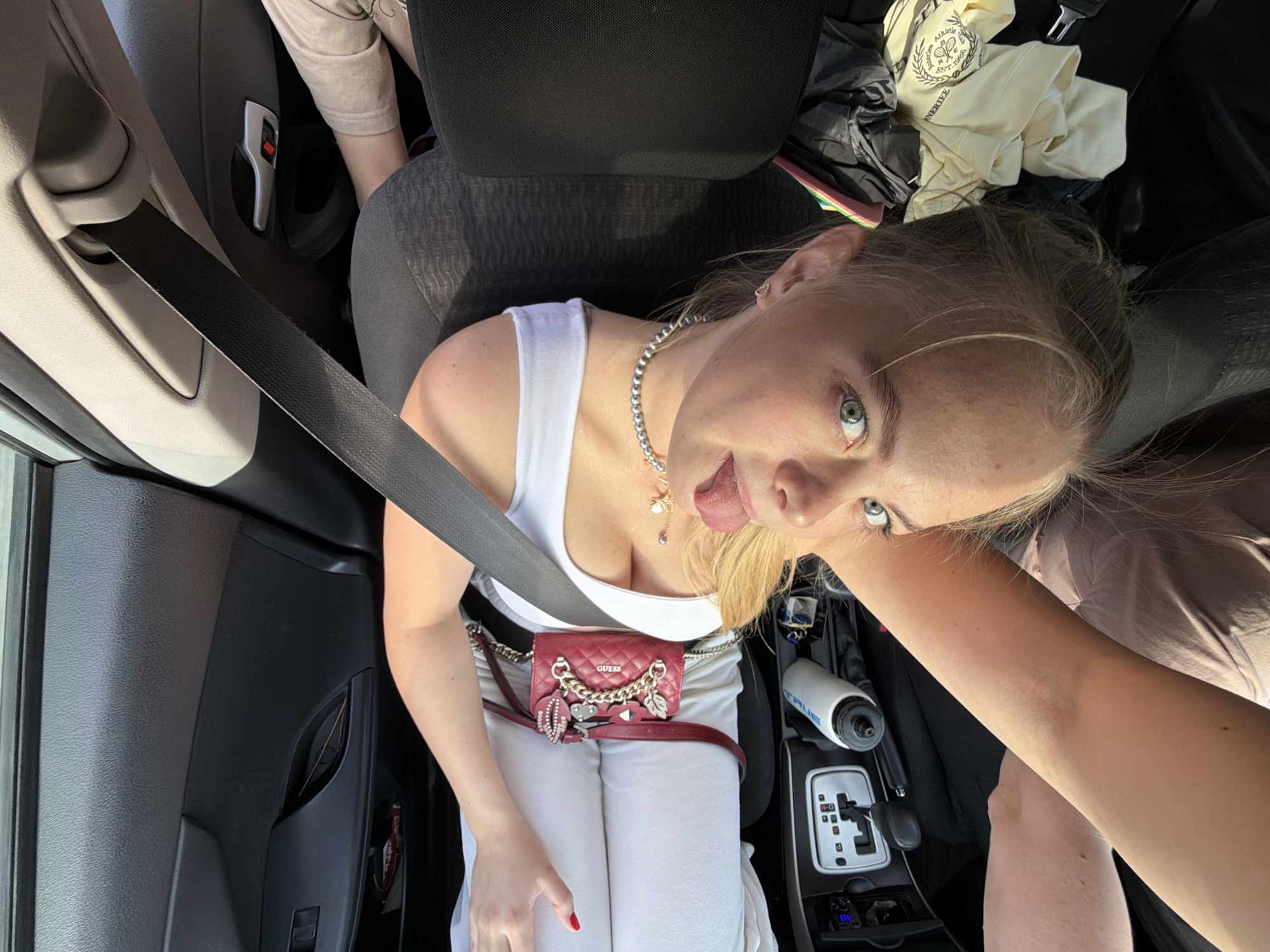 A young woman with blonde hair and blue eyes is sitting in the front seat of a car, wearing a white tank top and white pants, a silver necklace, and a red "Guess" belt bag. She is sticking her tongue out. She is wearing a seatbelt. A water bottle is next to the gear stick. Some clothing is on the back seat behind the headrest. The image was taken inside a car, but the location in the city of Izhevsk, Russia, is not discernible from the image.