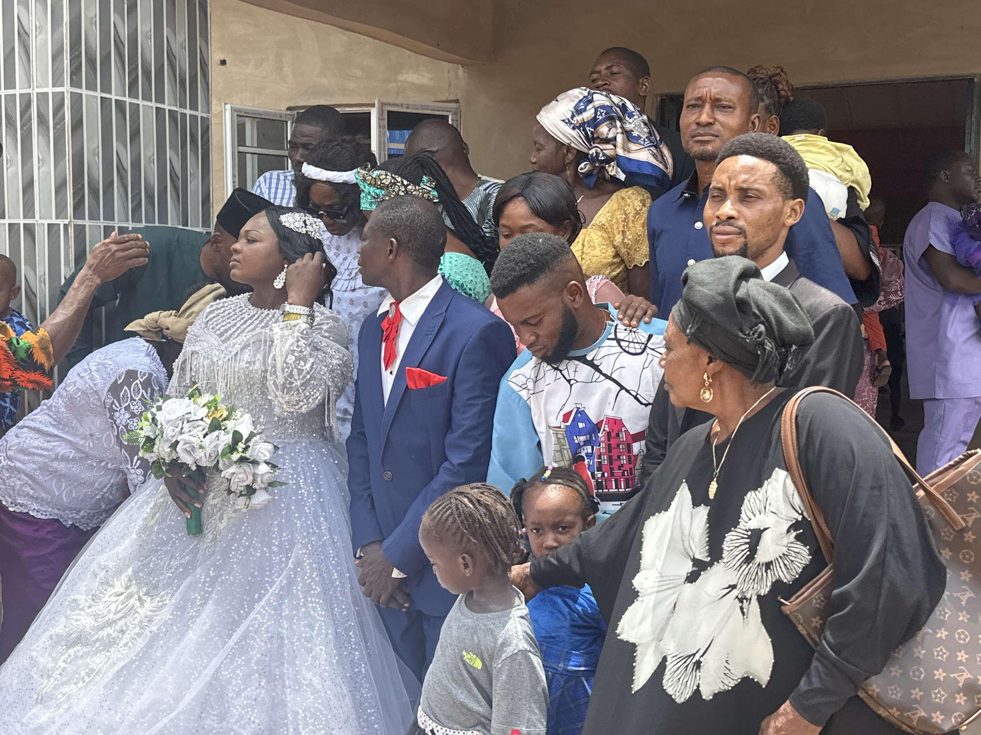 A wedding scene unfolds in an outdoor location in Unknown, Unknown, featuring a group of people gathered for the occasion. The bride, dressed in a sparkling white gown and holding a bouquet, stands beside the groom, who is wearing a blue suit with a red pocket square. Attendees are dressed in a variety of attire, including traditional headscarves, casual shirts, and suits. A child in a gray t-shirt is near the front, partially obscured by an adult wearing a black top with a white flower print and carrying a patterned brown bag. The event takes place near a building with a window.