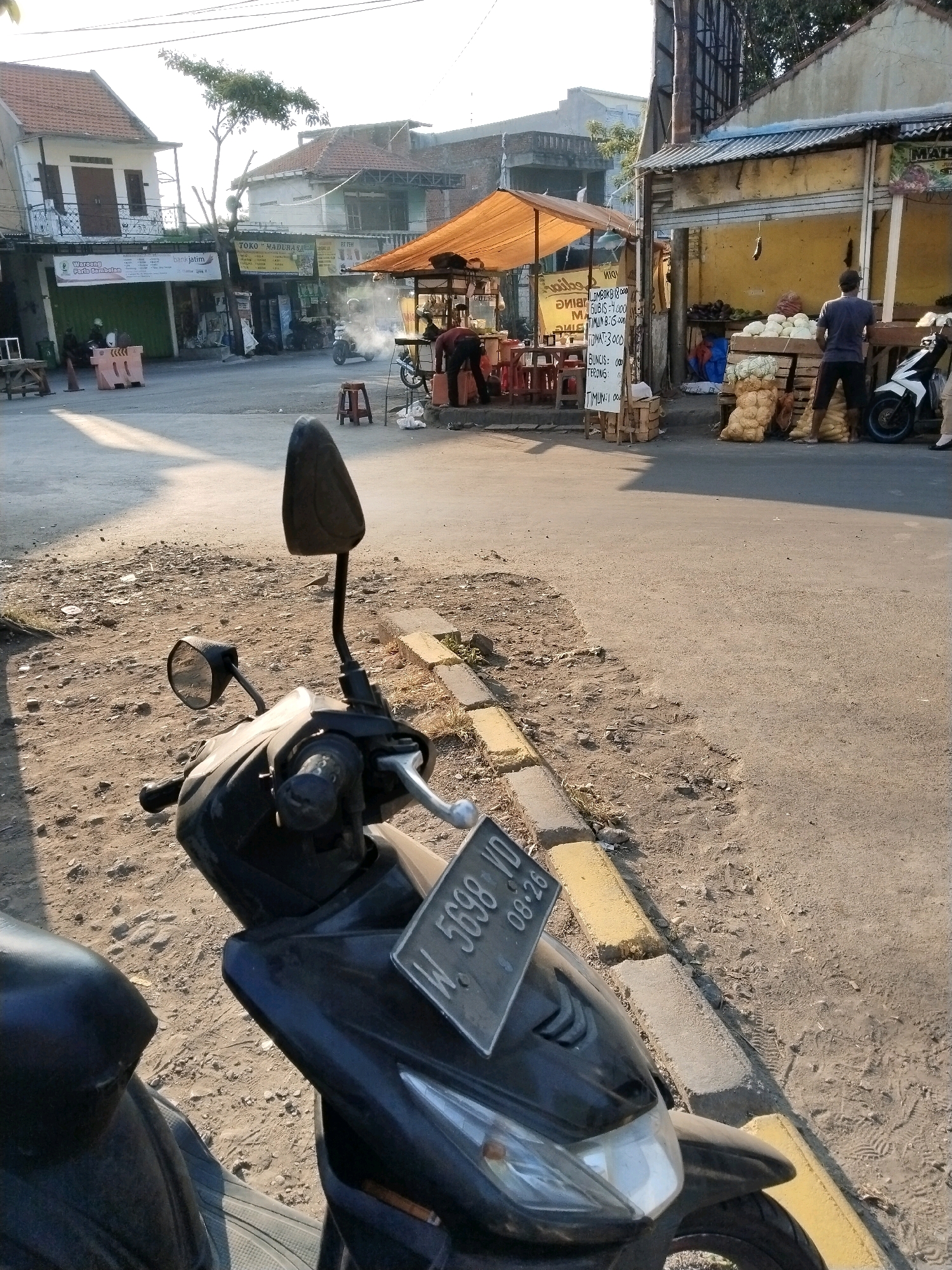 In the city of Wonocolo, Indonesia, a street scene unfolds under a clear sky. The foreground features a black motorcycle, its handlebars and license plate with the inscription "W 5698 VD 08-26" visible. The motorcycle is parked on a dusty, uneven surface with scattered debris and yellow-painted concrete curbs.
In the mid-ground, a street market is in full swing. A temporary food stall with an orange awning is set up, displaying tables and chairs. Several people are present, some appearing to be vendors and customers. One individual, wearing a blue shirt and dark shorts, stands by a stall laden with produce, including sacks of potatoes and what appear to be cabbages or similar vegetables. Another person in a dark shirt and pants is seen working behind a wooden counter. A white motorcycle is parked near the produce stall. Smoke rises from a small cooking area, suggesting food is being prepared.
The background reveals a row of buildings, some of which are shops or businesses. Signs are visible, including one that reads "TOKO MADURAJAYA". The architecture suggests a typical Indonesian urban setting with tiled roofs and a mix of single and multi-story structures. The overall impression is a lively, sun-drenched marketplace in a residential or commercial district.