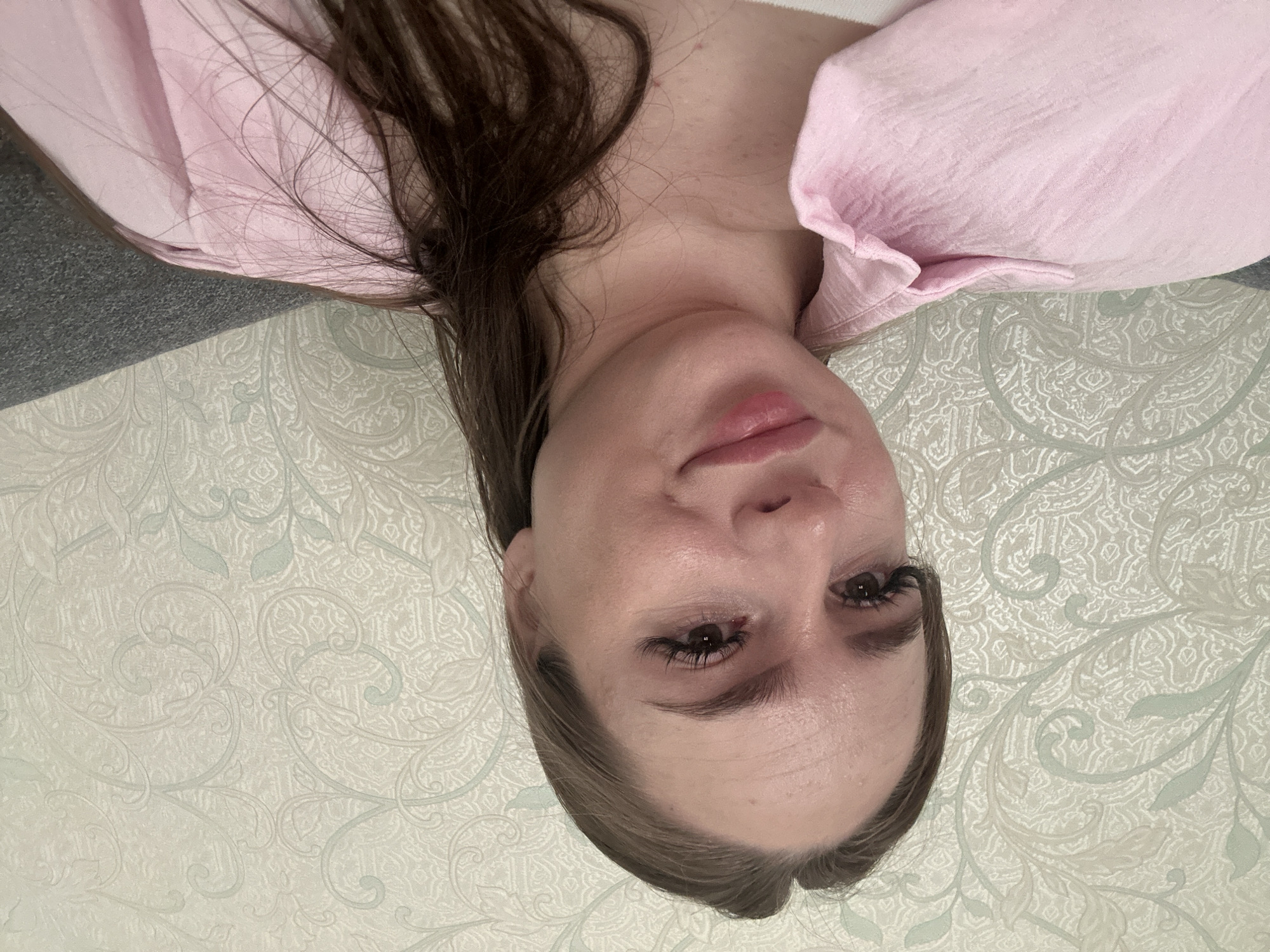 A young woman with long, dark brown hair is captured in a close-up, indoor shot. She is wearing a pink collared shirt, and her head is slightly tilted. The background features a textured wallpaper with an ornate, repeating pattern in pale green and off-white tones. The woman's face is the focal point, with her eyes looking towards the camera and a slight smile on her lips. Her makeup includes mascara and lip gloss. The lighting appears to be natural or soft indoor lighting, highlighting the texture of her skin and hair. The overall impression is a casual, personal portrait. The location is Unknown, Unknown.