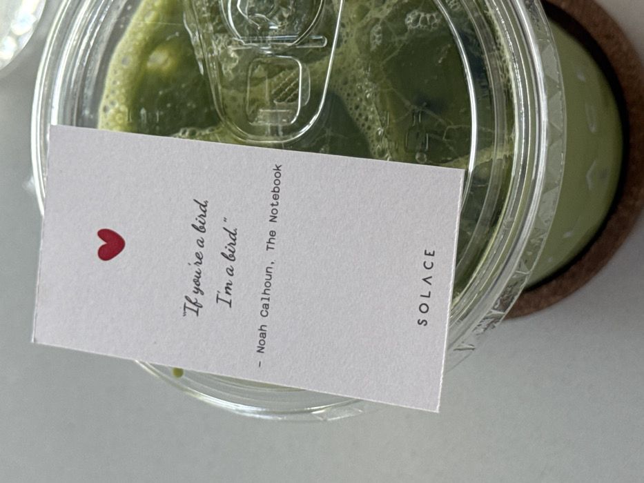 A clear plastic cup filled with a green beverage, possibly matcha, is displayed. A white card with a red heart sits on top of the cup. It features the quote "If you're a bird, I'm a bird." attributed to Noah Calhoun from "The Notebook." The word "SOLACE" is printed in simple, sans-serif font at the bottom right corner of the card. The cup rests on a brown coaster.