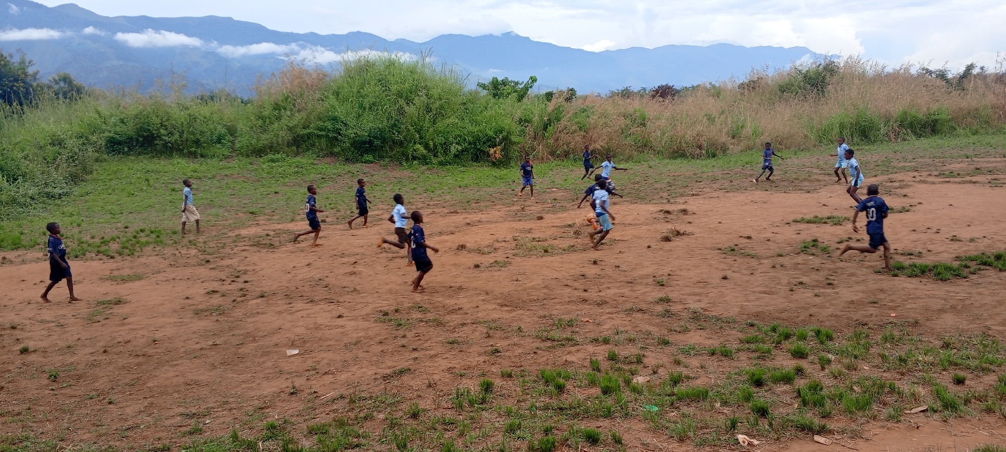 The image captures an outdoor scene in Ngalula, Democratic Republic of the Congo, featuring a group of approximately a dozen children, primarily boys, engaged in an informal game of soccer. The activity takes place on a reddish-brown dirt field, with several children playing barefoot. They are dressed in various light-colored shirts and shorts, predominantly blue and light blue, suggesting two loose teams or simply individual attire. One child on the far left wears a dark blue shirt with "QATAR AIRWAYS" visible, while another, seen from behind running towards the right, sports the number "10" on their dark blue jersey. A soccer ball is visible near the center, being kicked by one of the players.
The children are captured in various states of movement, running, kicking, and observing the play, conveying a sense of energetic and joyful engagement. The field is bordered by lush, untamed green and brown vegetation, including tall grasses and bushes. In the background, majestic mountains rise, their peaks partially obscured by soft, white clouds under a partly cloudy sky, creating a scenic and dramatic backdrop. The lighting suggests it is daytime, likely mid-day or afternoon, with bright but diffused light. The overall scene depicts vibrant childhood play in a natural, rural setting.