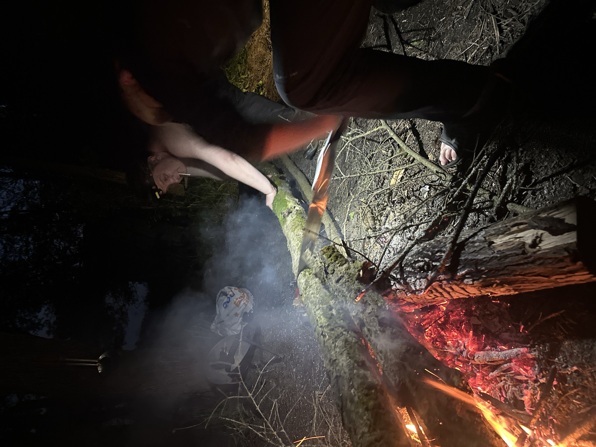 The image depicts an outdoor nighttime scene near Юзуфоўскі сельскі Савет, Belarus, dominated by a large bonfire. A fallen tree trunk with moss on its bark serves as a makeshift support structure for the fire. Flames are visible emanating from the burning wood and embers, producing smoke. A person is positioned near the fire, seemingly tending to it. Another individual, shirtless and smoking, is positioned on the other side of the tree trunk. A bag, partially obscured by smoke, rests on the ground nearby. The surrounding environment appears to be a wooded area, with branches and foliage visible in the background.
