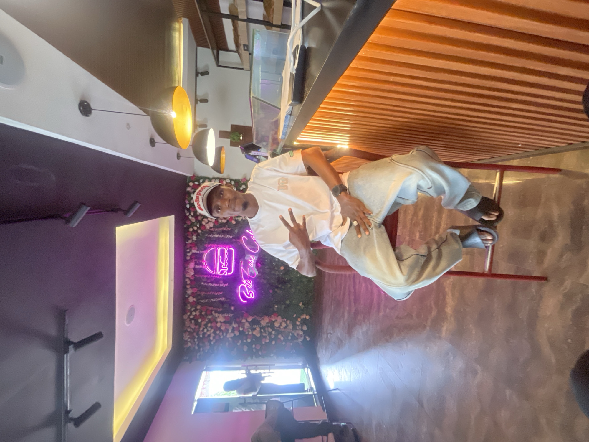 A man wearing a white t-shirt, light-colored pants, and a hat is sitting on a red chair inside what appears to be a restaurant in Adankolo, Nigeria. He is making a peace sign with his hand. Behind him is a wall decorated with flowers and a neon sign that reads "Bite Bake Cafe." The restaurant has a modern design with pendant lights, a counter with wooden slats, and a view of the outside. A person and some furniture are visible through the window.