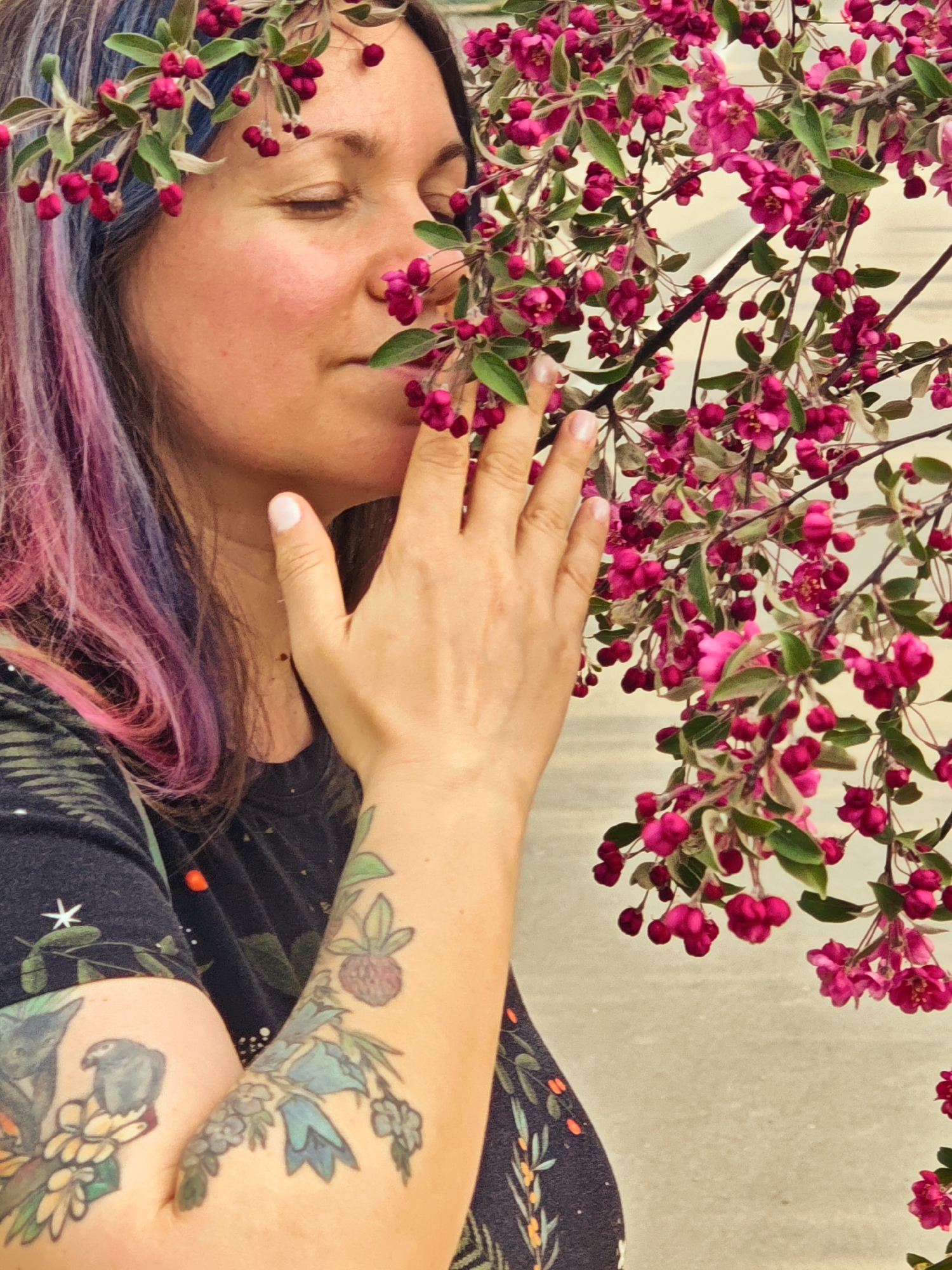 A medium shot depicts a woman with her eyes closed, her face partially obscured by a branch laden with small, bright pink flowers. She appears to be enjoying the scent of the blossoms, her hand raised as if to protect the delicate blooms. She is wearing a dark, floral-patterned shirt and has vibrant pink and blue streaks in her dark hair, along with a floral crown. Her arms are adorned with colorful botanical-themed tattoos. The background appears to be a neutral, slightly textured surface. The location is not specified.