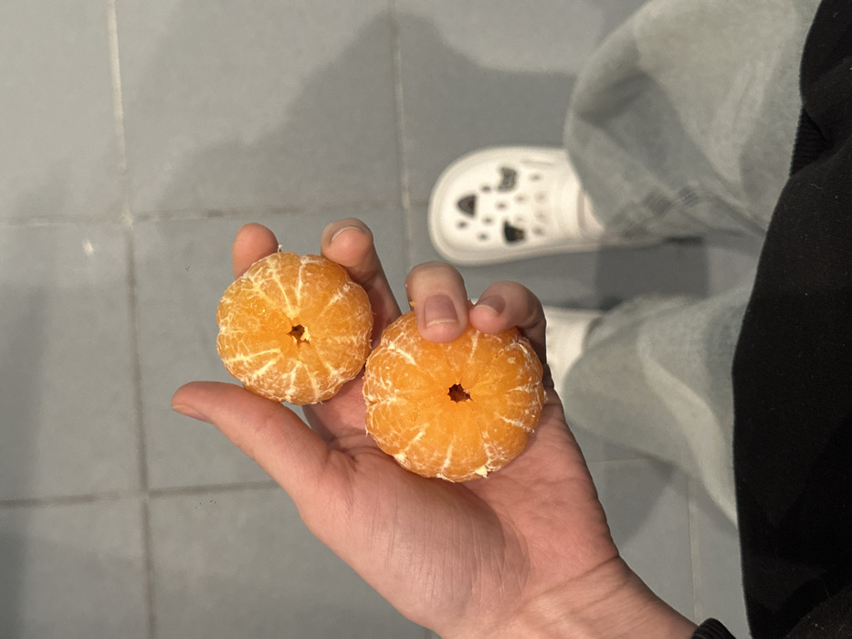 A hand holds two peeled tangerines, each displaying a visible hollow at the top. The skin is segmented, exposing bright orange flesh. The person is wearing light-washed jeans and a black shirt, with a white Croc shoe with black charms visible in the background. The setting appears to be indoors with grey tiled flooring.