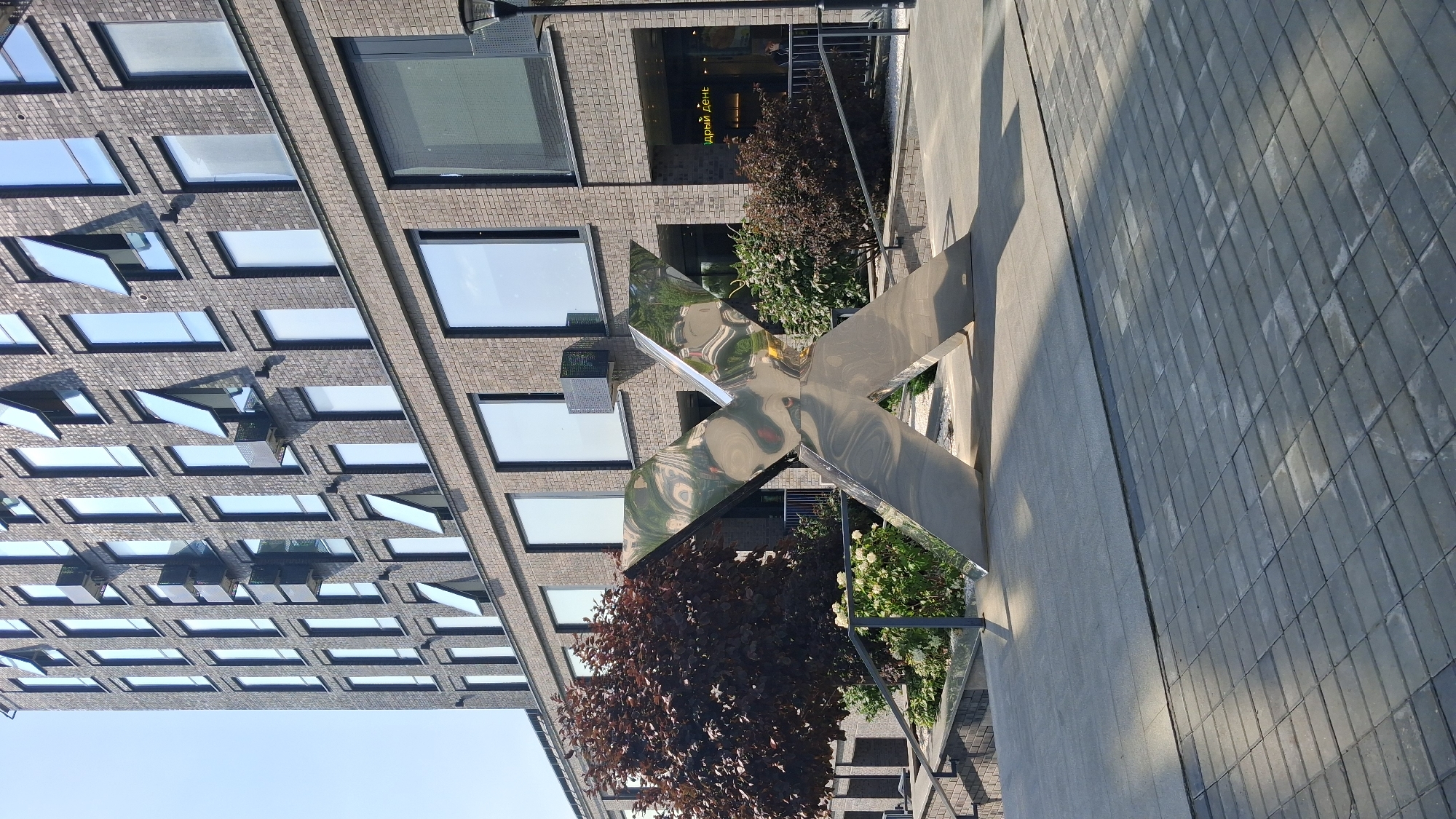 A polished, metallic sculpture, shaped like an abstract "X", stands prominently in an urban outdoor setting. The sculpture is silver and reflects the surrounding environment, including the sky, building, and greenery. It is positioned on a paved surface made of gray rectangular stones.
Behind the sculpture, a multi-story modern building with a dark brick facade rises. The building features numerous windows, many of which are large and rectangular, with some having hinged sections open. Visible on the building's facade are elements like air conditioning units and balconies.
To the left of the sculpture, there is a dense, dark red-leafed tree and a planter with white flowers and green foliage. A metal railing and steps are also visible leading up to the building.
In the background, beyond the sculpture and to the right, a store entrance is visible with signage. A lamppost is positioned near the building's entrance. The overall scene suggests a public space or a commercial area in a city. The image appears to be taken during the daytime, with sunlight casting shadows on the ground.
The location is in Novosibirsk, Russia, specifically at the Novosibirsk State University of Economics and Management (NSUEM). The sculpture is a well-known landmark outside the university.