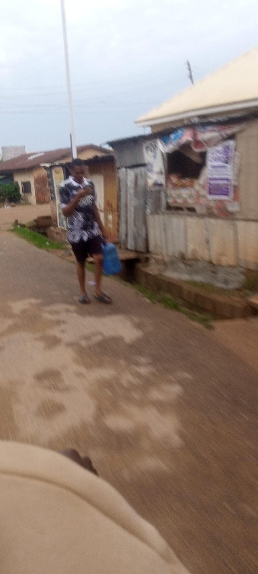 A man walks on a narrow road in Narayi, Nigeria. He wears a patterned short-sleeved shirt, shorts, and sandals, and carries a blue plastic bag. The road is paved but stained, with a narrow ditch alongside. Buildings, including shops with corrugated metal, line the road. The sky is overcast.