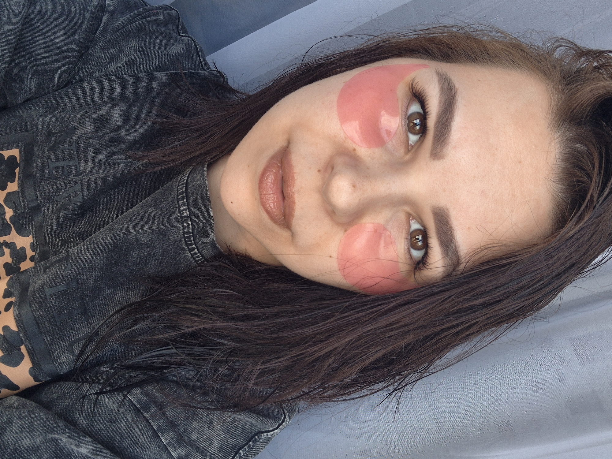 A close-up, high-angle portrait features a young woman with long, dark brown hair and warm brown eyes, located in Tobolsk, Russia. She is shown applying pink, translucent under-eye patches to her face, one beneath each eye, indicating a self-care or beauty regimen. Her eyebrows are neatly groomed, and she wears a subtle, natural-toned lipstick.
The woman is dressed in a dark, possibly acid-washed or distressed, denim-like top or jacket. Visible on the left side of her garment is a brown and black leopard or cheetah print pattern, along with faded black text that clearly shows the word "NEW" and partial letters suggesting other words. Her expression is calm and relaxed, with a hint of a gentle smile. The background consists of a soft, light grey or blue textured fabric, possibly a sheet or wall covering, and the lighting is bright and natural, suggesting daytime. The image is captured with a diagonal tilt, emphasizing the woman's face and eye area.