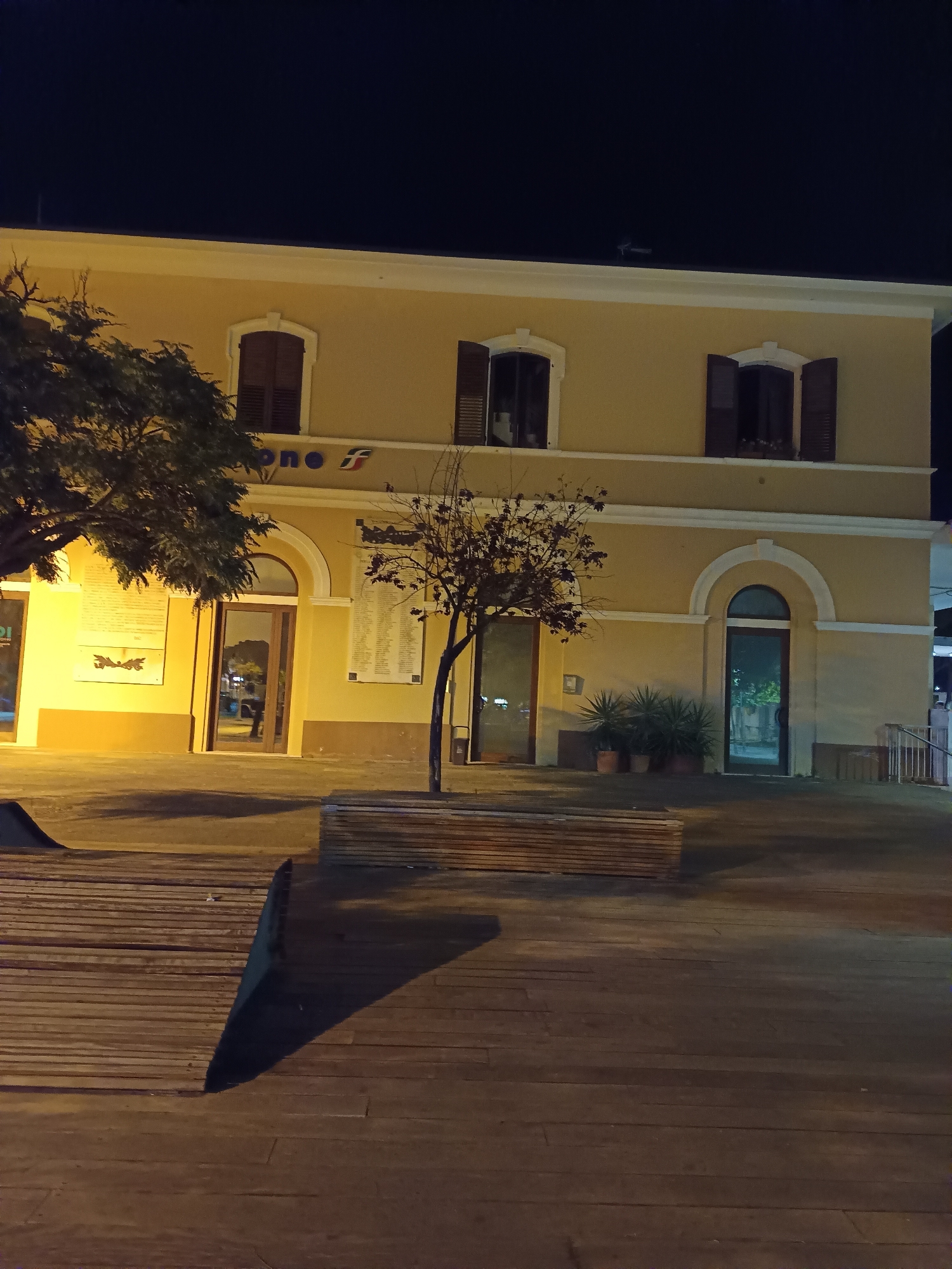 The image shows a building, likely a train station, at night in Roseto degli Abruzzi, Italy. The building is a two-story structure painted yellow with dark brown shutters on the windows. There are arched doorways and windows. A sign with the letters "uno f" is visible above one of the doorways. In front of the building is a paved area with wooden benches. There are trees and potted plants around the building. The lighting suggests artificial illumination.