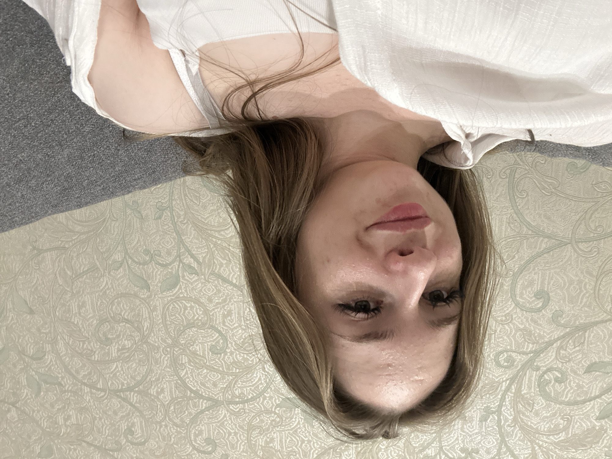 A young woman with long brown hair is captured in a close-up, high-angle shot. She is wearing a white tank top and an unbuttoned white shirt. Her head is tilted slightly to her left, and she is looking directly at the camera with a faint smile. The background is a patterned wallpaper. The image appears to be taken indoors, possibly on a couch, given the presence of a gray textured surface visible on the right side of the frame. There is evidence of makeup, including mascara and lipstick. Her forehead shows some slight blemishes or oiliness. The lighting suggests an indoor environment, possibly natural light or ambient room light.