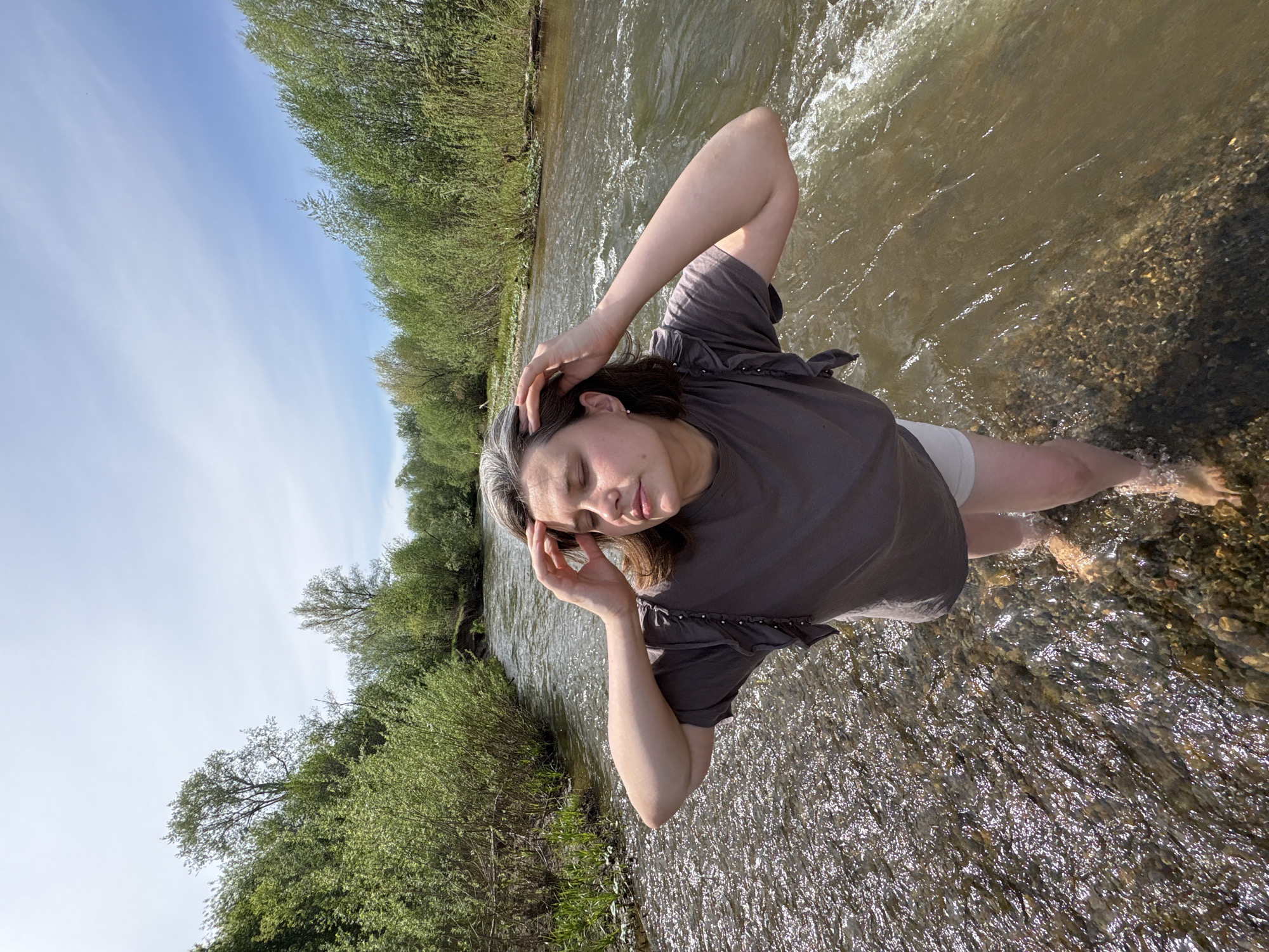 A woman stands in a shallow, rocky river, likely near Nizhnyaya Chermoda, Russia, given the green deciduous trees lining the riverbank and the regional sunlight. She is wearing a gray t-shirt and white shorts. Her graying hair is pulled back, and her eyes are closed. Both hands are touching her head. The river's water is relatively clear, allowing the rocky riverbed to be visible. The sky is blue with scattered wisps of clouds.
