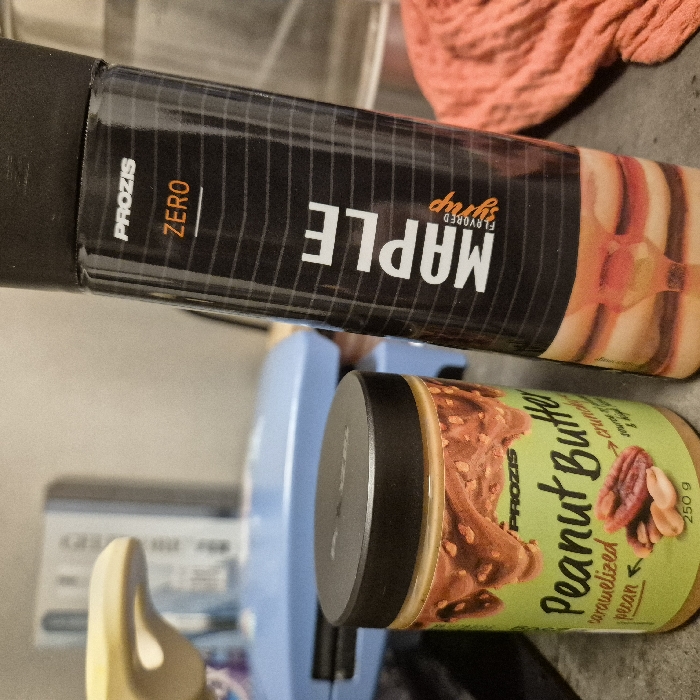Two branded food products are displayed on a dark, textured surface within an indoor environment in Strasbourg, France. On the left, a glass jar with a black lid contains "Prozis Peanut Butter caramelized pecan," a 250g product featuring a green and yellow label with graphics of pecans, peanuts, and caramel drizzle. To its right, a tall, black bottle with a black cap, labeled "Prozis ZERO Maple Flavored Syrup," stands upright, its label depicting pancakes with syrup. The background, out of focus, shows a light blue object, likely an appliance, and a crumpled reddish-orange cloth positioned to the right.