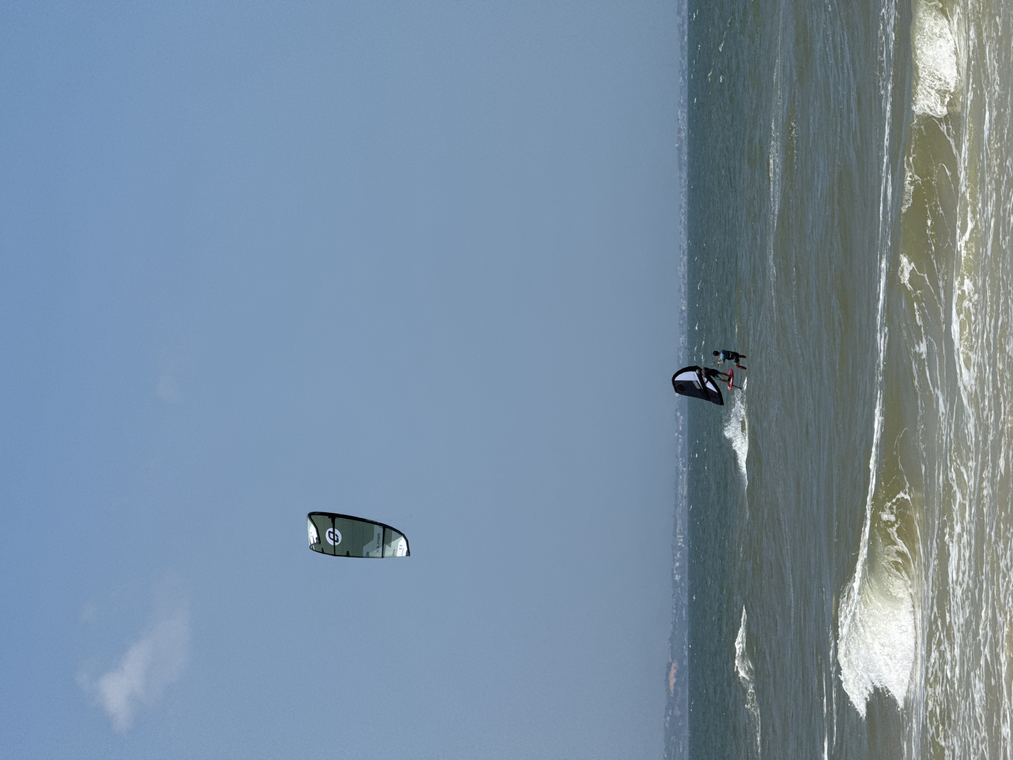 The image depicts a bright, sunny day at a beach in Mũi Né, Vietnam. The sea is a deep blue-green, with white-capped waves rolling towards the shore. In the distance, the coastline is visible, lined with low-lying buildings.
The main subjects in the image are two individuals engaged in water sports. One person is kiteboarding, with a large, green and white kite visible high in the clear blue sky. The kite has a circular logo with a stylized "e" or "3" inside. The other person is further out on the water, windfoiling. This individual is on a board with a hydrofoil underneath, and they are using a sail that is black and white with a distinctive curved shape. This windfoiler is wearing a dark blue top and appears to be actively engaged in the sport.
The foreground shows the churning surf and the sandy beach, although the beach itself is not the primary focus. The overall impression is one of an active and enjoyable day at the seaside, with the kites and sails adding dynamic elements to the scene against the backdrop of the ocean and the distant town.