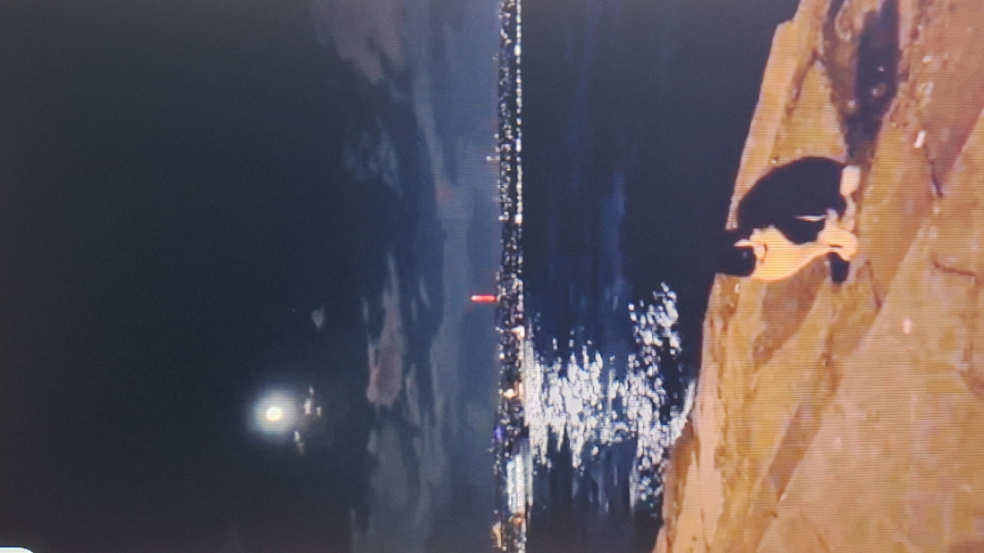 The image displays a serene nighttime scene, likely captured from a waterfront embankment or pier. In the foreground, a distinct black and white cat is perched on a light-colored, possibly rocky or concrete surface, its head turned downwards as if observing something in the water below.
The body of water reflects numerous bright lights from a distant cityscape, creating shimmering streaks across its dark surface. A prominent feature in the distant urban panorama is a tall, slender red light source, possibly a lighthouse or a beacon, standing out against the smaller city lights. The horizon is marked by a continuous line of these urban lights.
Above the distant city and water, the night sky is dark, with visible cloud formations. A bright, circular light source, likely the moon, is faintly visible through a patch of clouds in the upper left portion of the image, casting a soft glow. The overall setting suggests a coastal or lakeside urban environment at night. No specific location cues are identifiable, and there is no visible text.