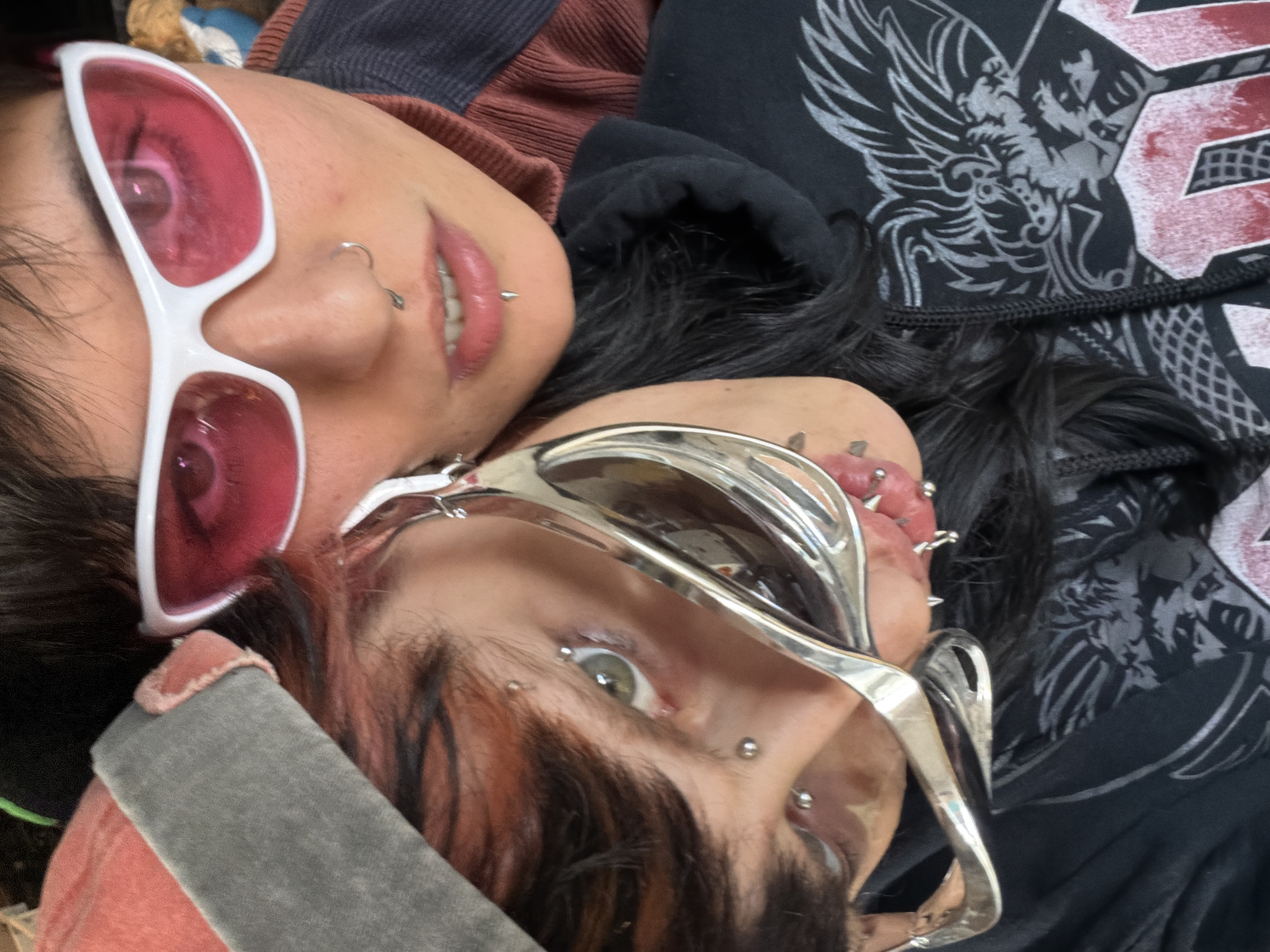 Two individuals are captured in a close-up shot. The person on the upper left wears white-framed glasses with pink lenses and has piercings on their nose and lip. The other person, positioned at the lower right, is wearing silver, futuristic-looking eyewear and has multiple piercings around their lips and brow. Their hair is dark with reddish-brown streaks. The background shows glimpses of clothing: a brown top on the upper left, and a black hooded sweatshirt with a gray design on the upper right. The image is close-up, providing a detailed view of the individuals' faces and accessories. The city of Sofia, Bulgaria is not discernible in the image.