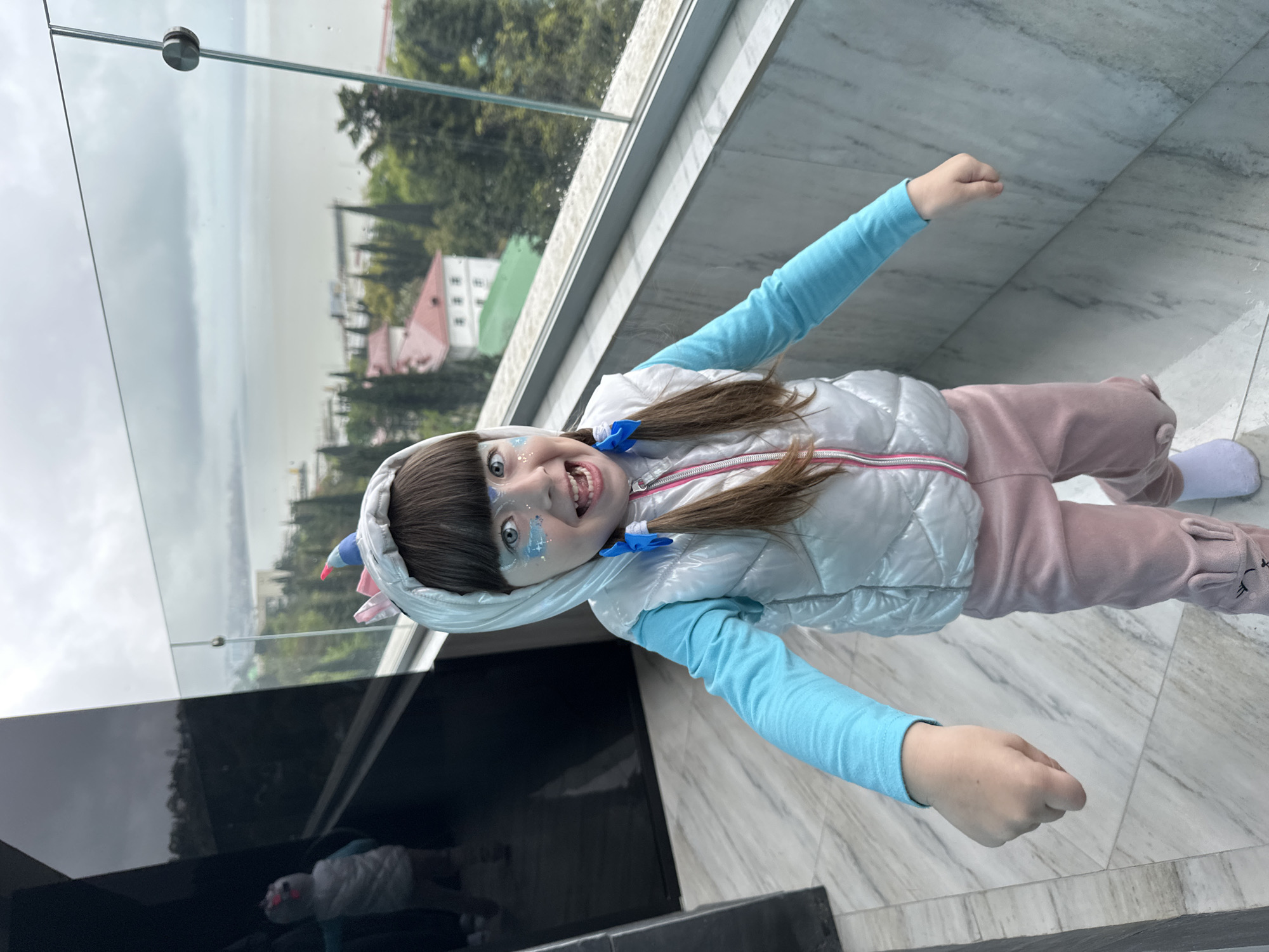 A young girl stands on a marble floor next to a black structure, possibly a counter or wall, near a balcony railing of transparent glass. She is wearing a light blue long-sleeved shirt, a white sleeveless quilted vest with pink trim, and light pink sweatpants with animal ear details on the lower legs. She also wears a hood with a unicorn horn attached. The girl has brown hair styled in pigtails with blue ribbons and blue glitter or face paint on her face. She is smiling with her mouth open and arms outstretched.
Behind the glass railing, a view of Sochi, Russia is visible, featuring green trees, buildings with red roofs, and a hazy sky or sea in the distance.