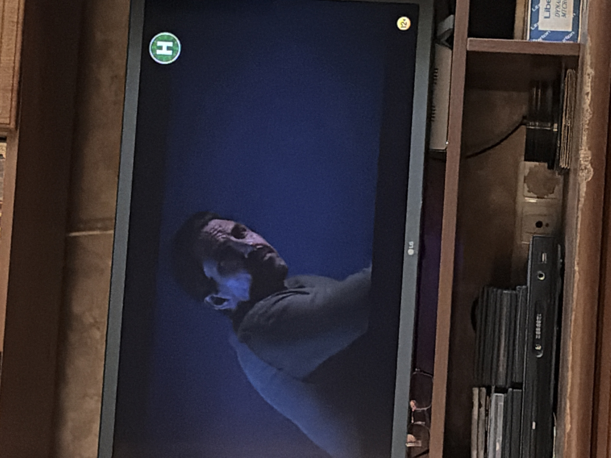 The image is a medium shot of a man's face and upper body displayed on a television screen. The man is looking to his right and appears to be in his 40s or 50s, with a receding hairline and a mustache. He is wearing a dark-colored sweater. The television screen is dark blue with a faint blue light source from the left. In the upper right corner of the screen, there is a green circle with a white "H" inside. In the lower right corner of the screen, there is a yellow circle with "12+" inside. The television is placed on a wooden shelf unit. Below the television, on the shelf, are several stacked items, including what appears to be a DVD player or VCR, and a stack of DVDs or CDs. There is also a small stack of books or boxes to the right. The overall setting appears to be a home entertainment area.