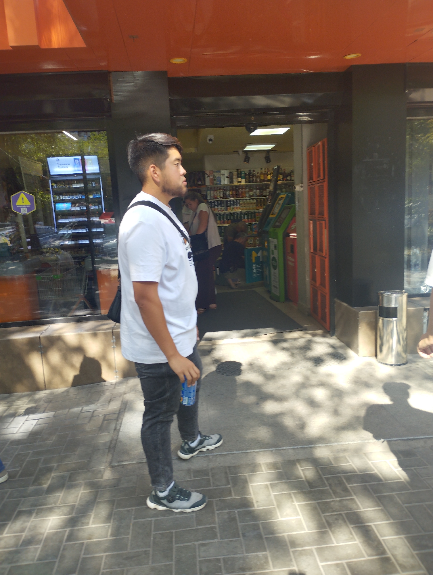 A man stands on a sidewalk in front of a store with a black facade. He is wearing a white t-shirt, dark jeans, and athletic shoes. He is holding a blue can in his right hand. In the background, inside the store, shelves of products are visible, including bottles of alcohol. There are also gaming machines in the store. The sidewalk is made of brick pavers, and shadows of trees are cast on the ground.