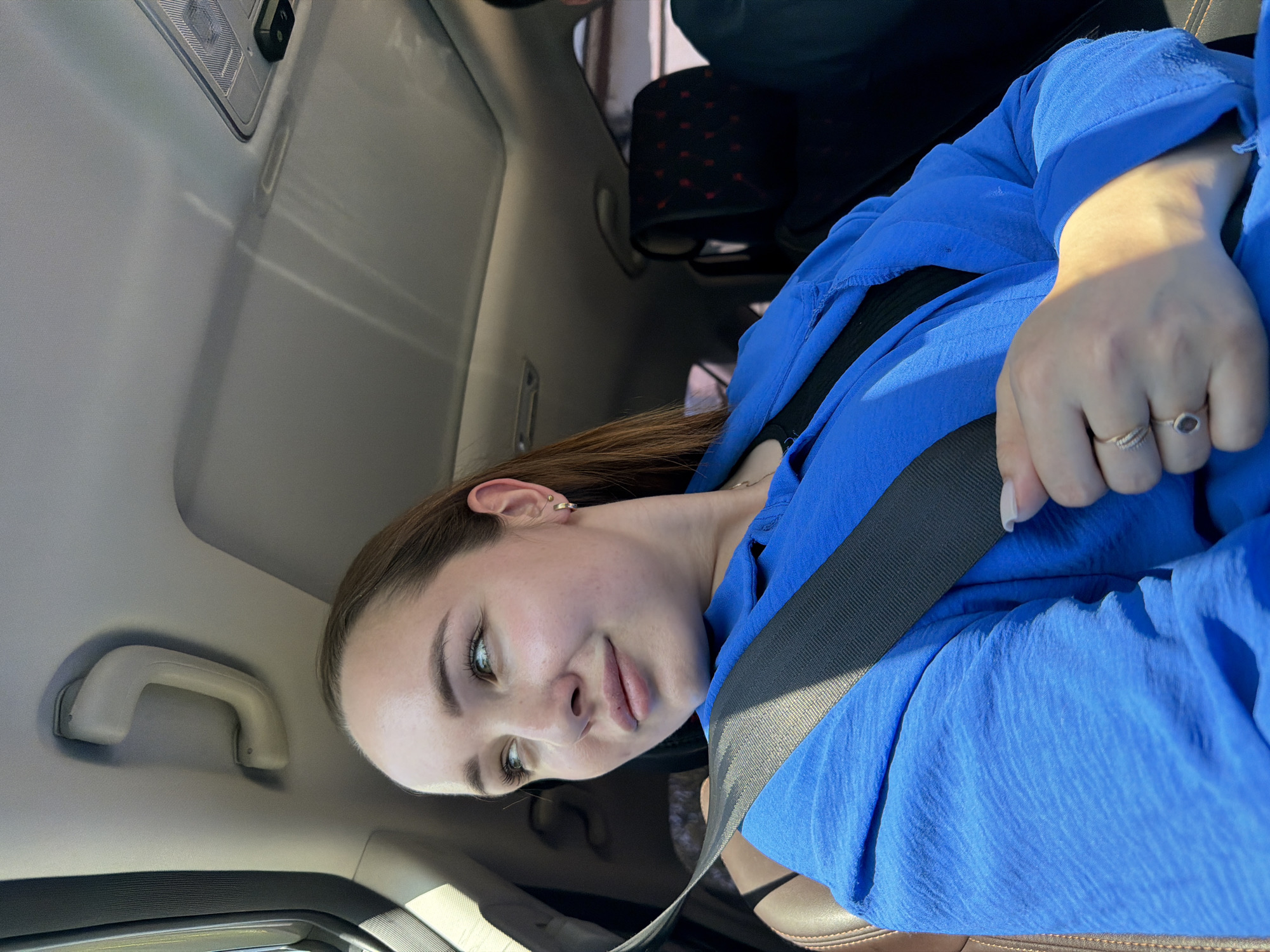 A young woman sits in a car, wearing a blue garment and a seatbelt. The interior of the car is visible, including the ceiling, a passenger seat, and a grab handle. The city of Chaikovsky, Russia, cannot be determined from this image.